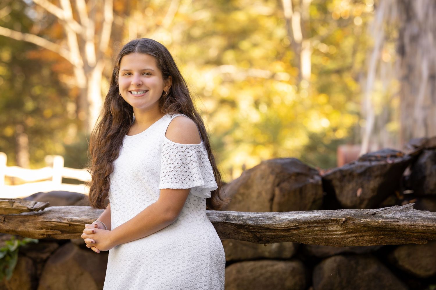 A young girl in a white lace dress poses outdoors against a natural backdrop with golden sunlight filtering through trees.