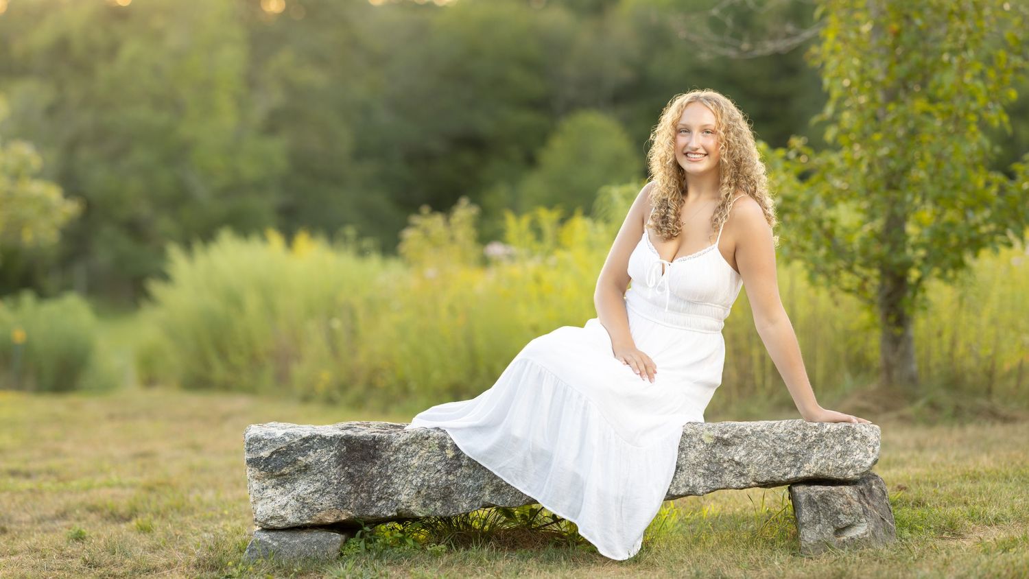 A bride in a white wedding dress poses gracefully on a rustic stone wall surrounded by lush green foliage.