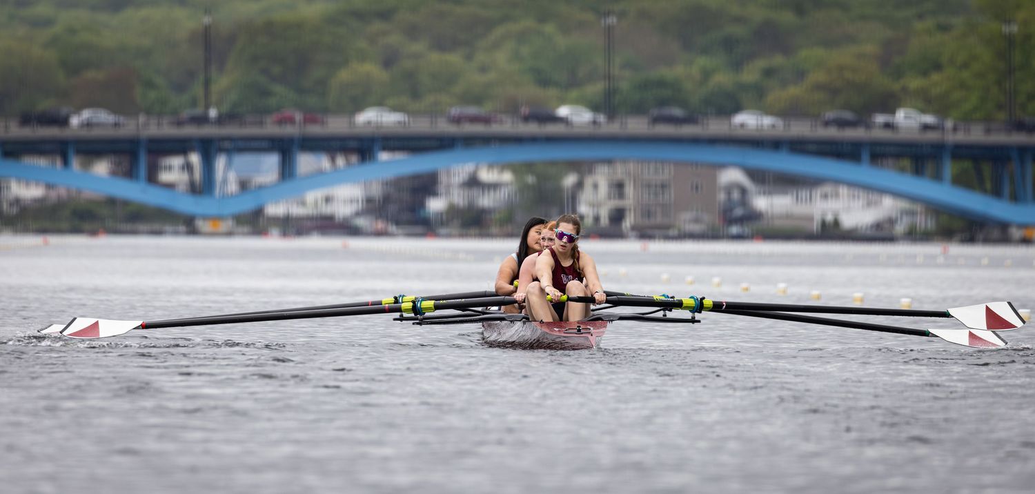 Two rowers glide across calm waters in a racing shell with a blue bridge visible in the background.