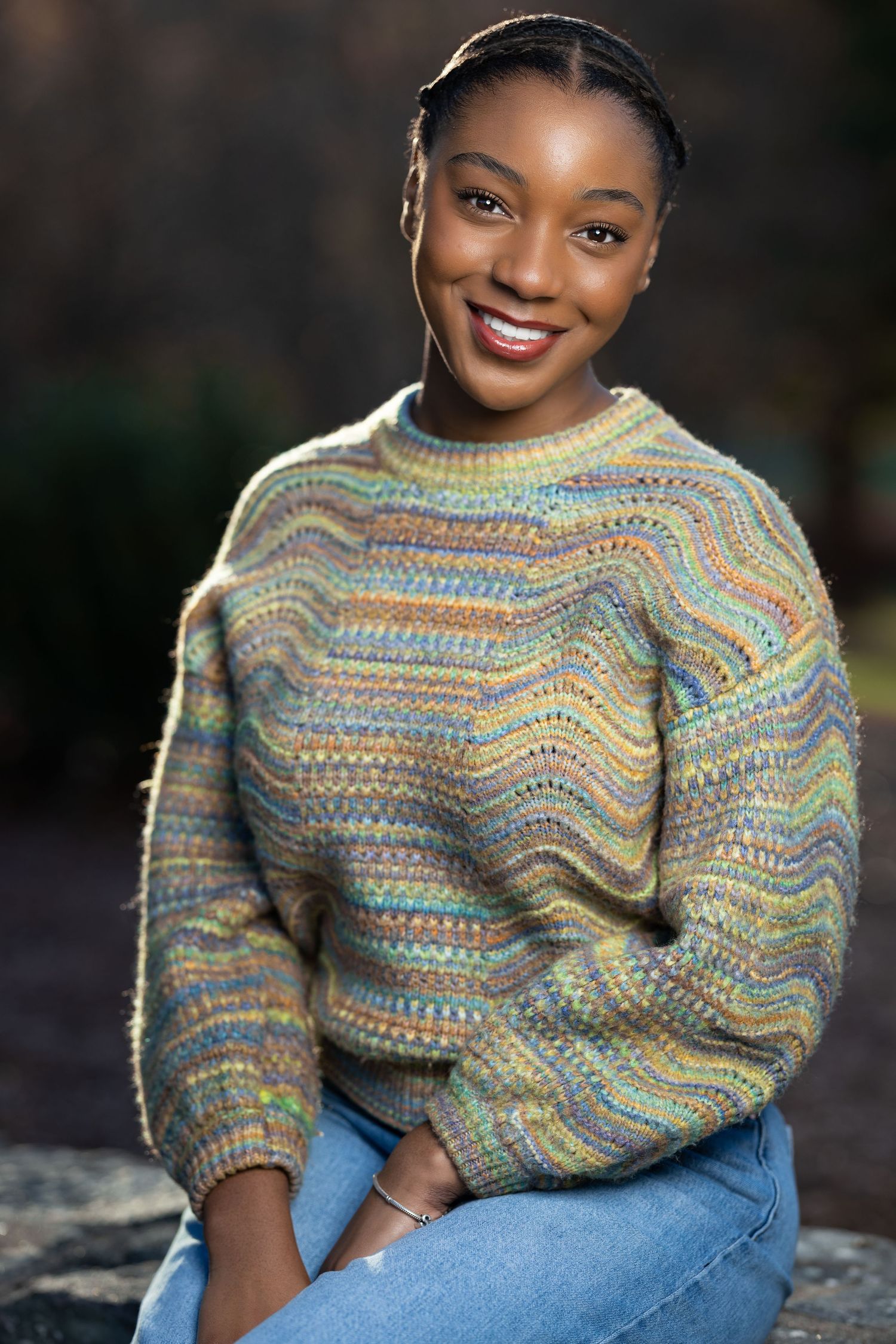Portrait of someone wearing a colorful patterned knit sweater and blue jeans while sitting outdoors.