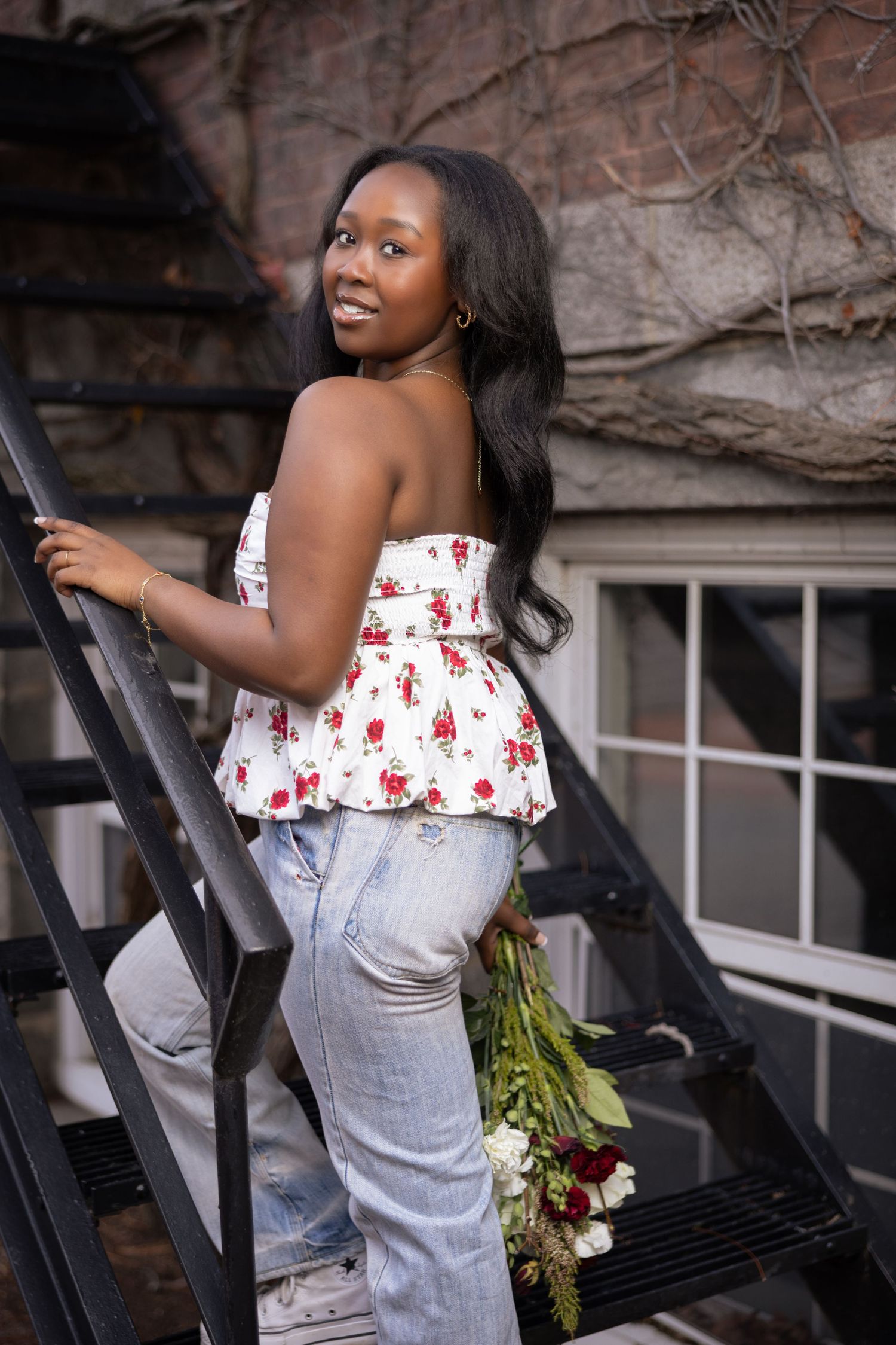 Woman in floral peplum top and jeans poses on metal fire escape stairs with flowers.