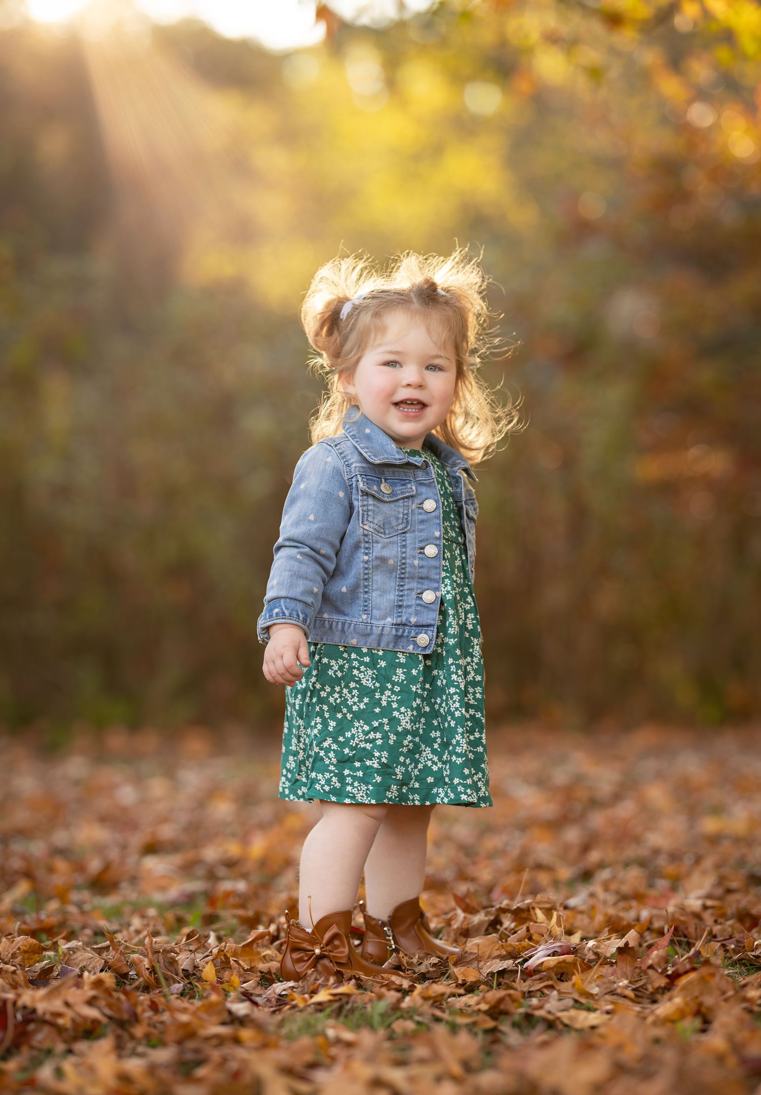 A child in a denim jacket and floral dress plays joyfully among fallen autumn leaves during golden hour.
