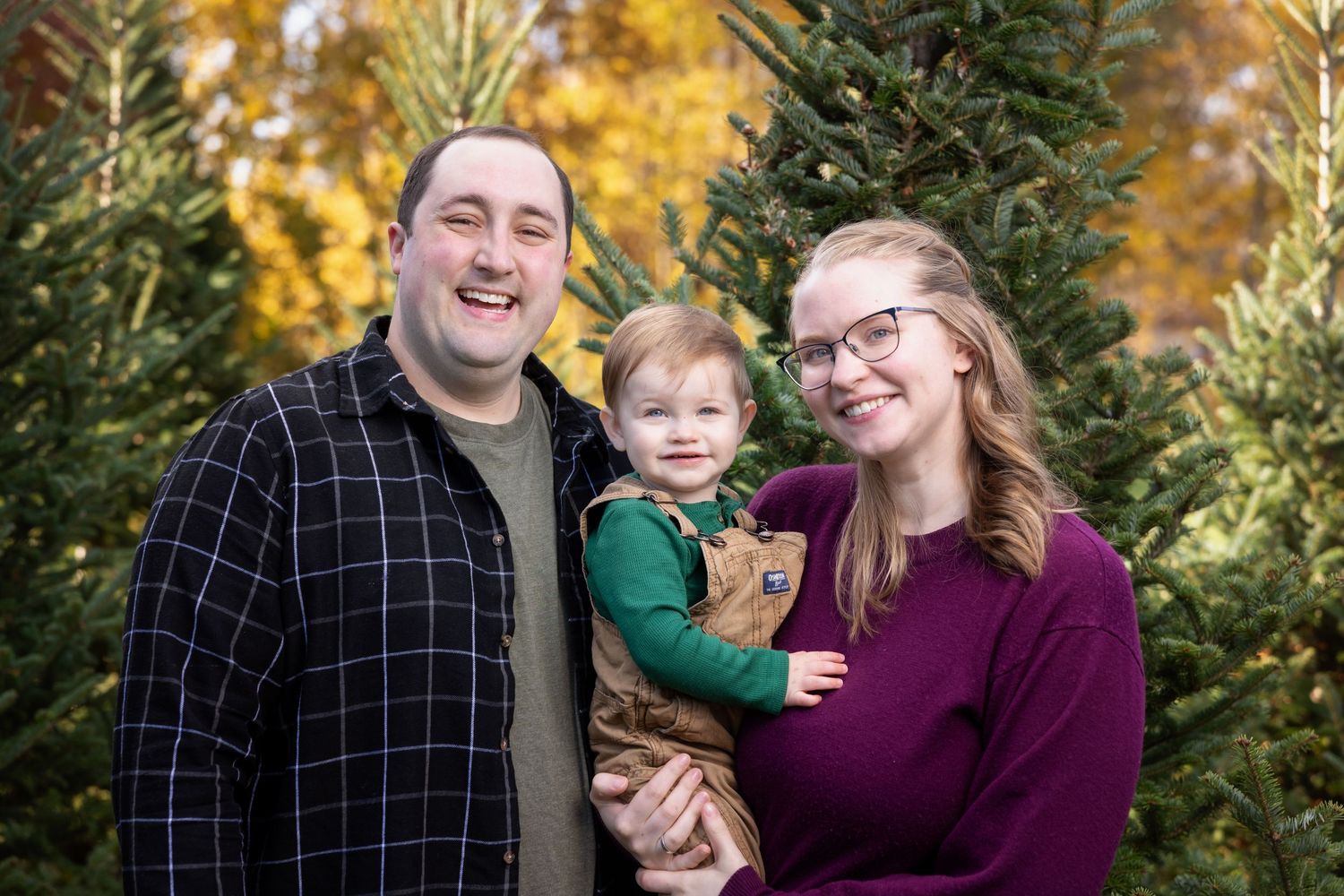 A family wearing plaid and purple poses together among autumn trees with golden foliage in the background.