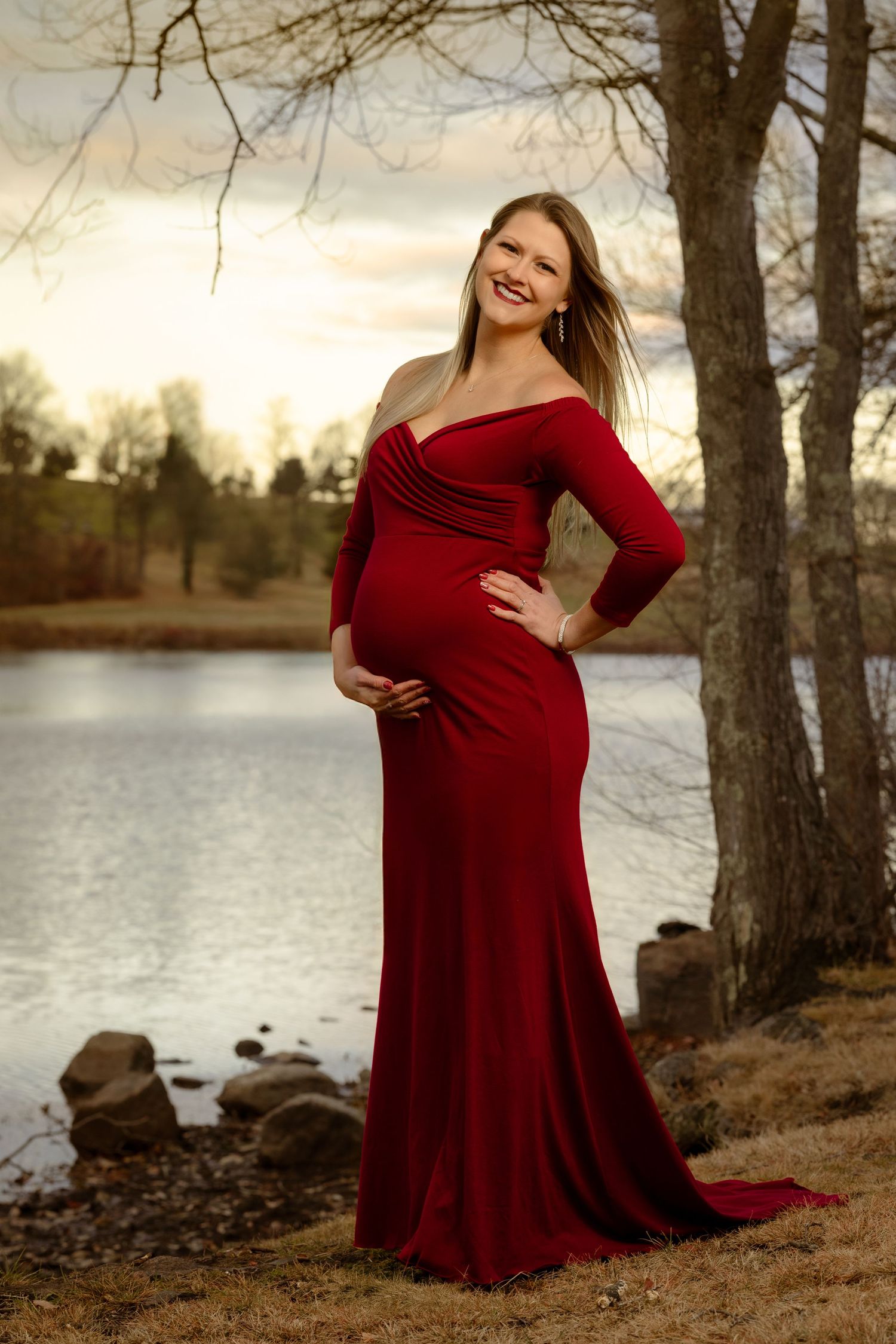 A flowing red maternity dress is showcased in a natural lakeside setting during golden hour with trees in the background.