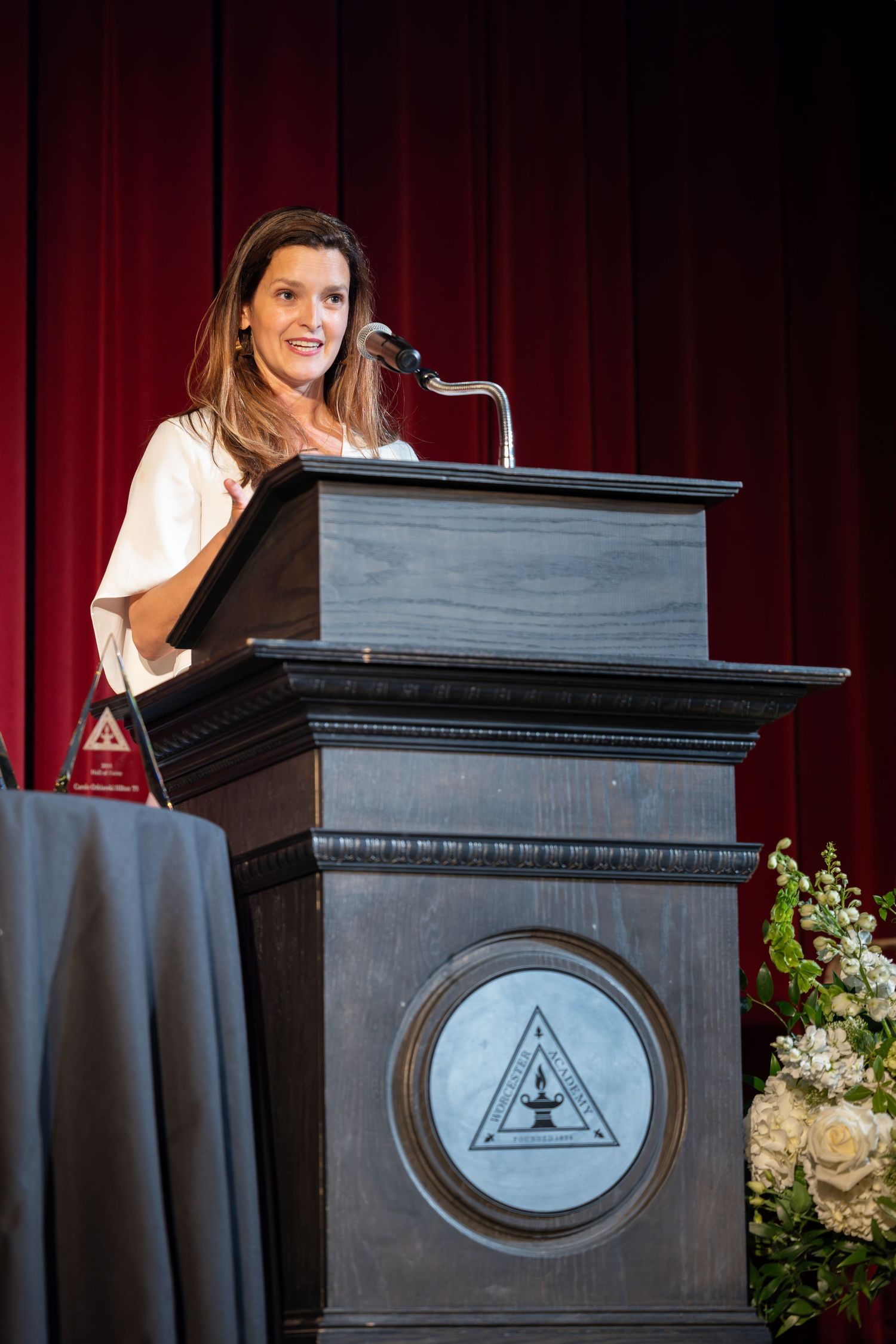 Speaker stands at wooden podium with triangular emblem and floral arrangements on stage with red curtain backdrop.