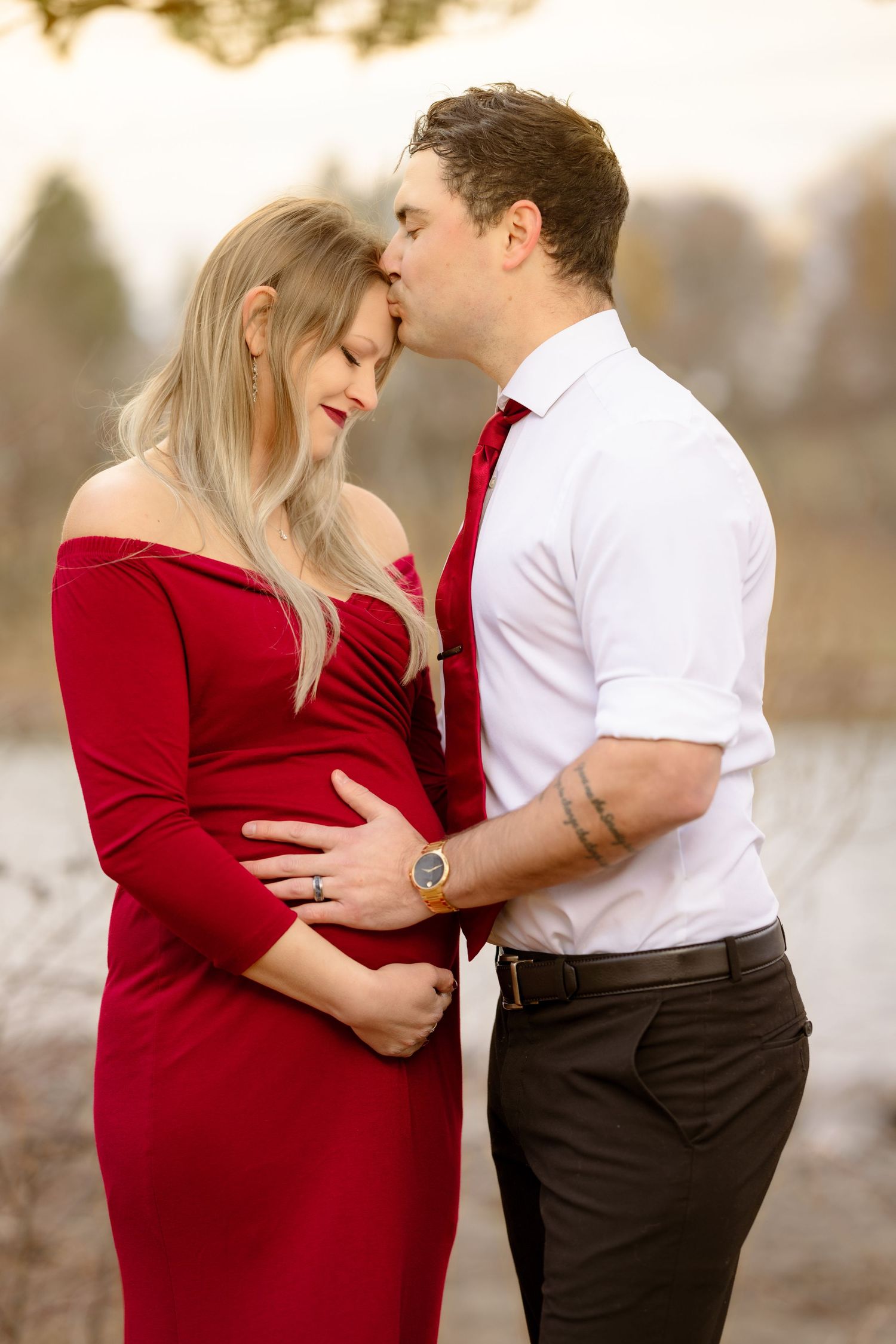 A couple shares an intimate moment outdoors, with her in a red dress and him in a white shirt and red tie.