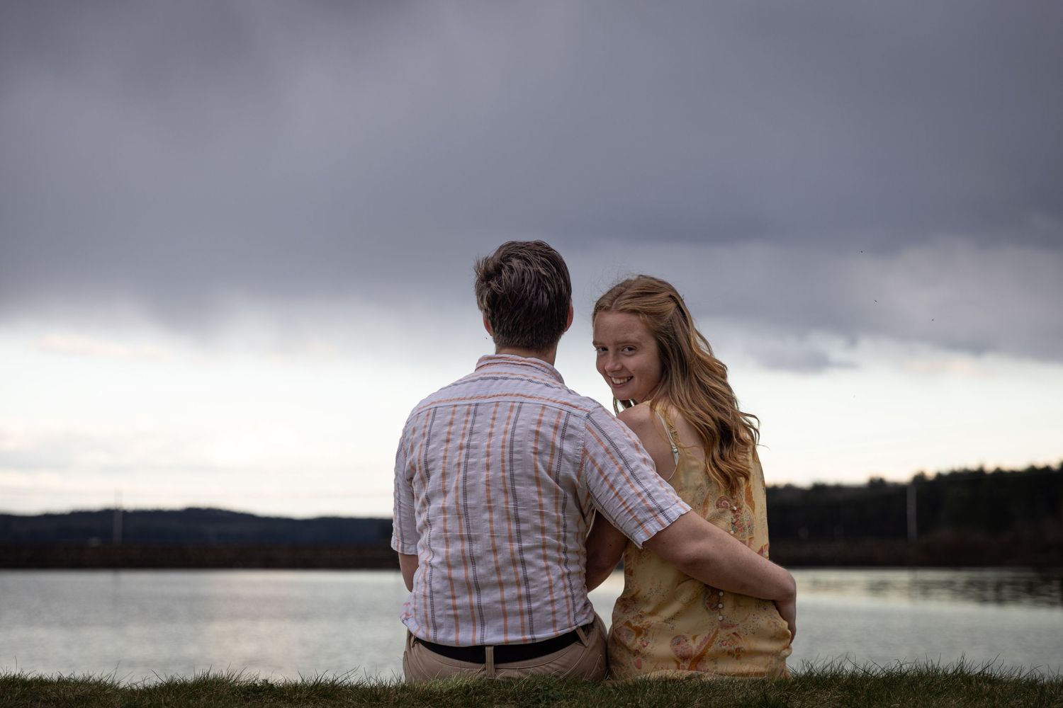 A couple sits together on grass overlooking a lake with stormy clouds gathering in the background.
