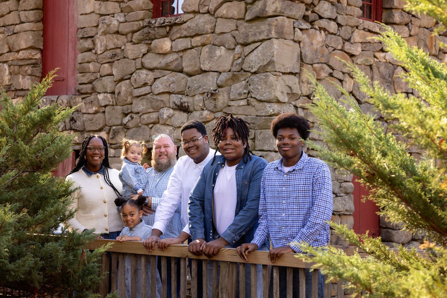 A group stands together smiling in front of a rustic stone wall with evergreen shrubs in the foreground.