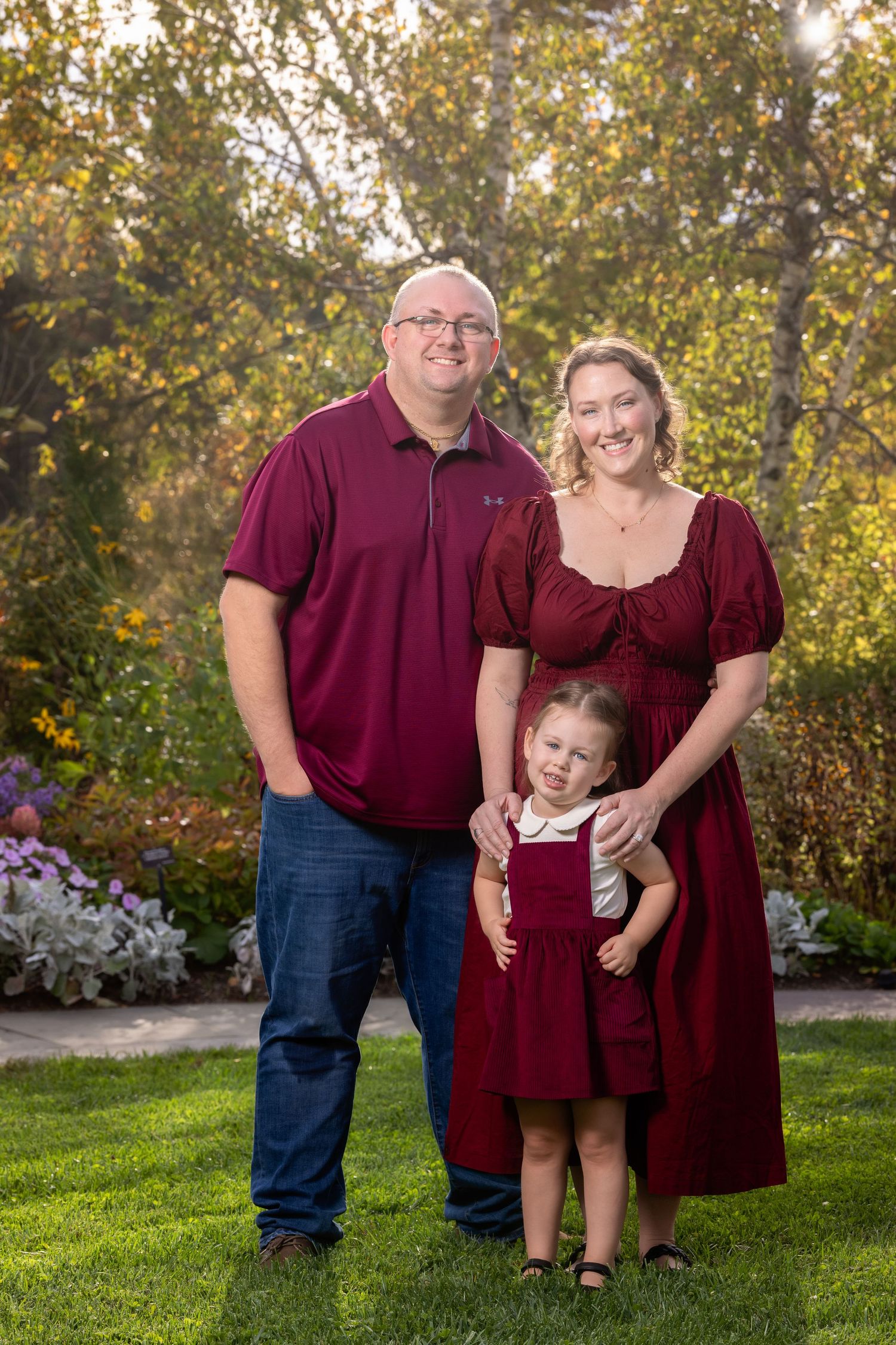 A family of three wearing coordinating burgundy outfits poses together outdoors in front of autumn foliage.