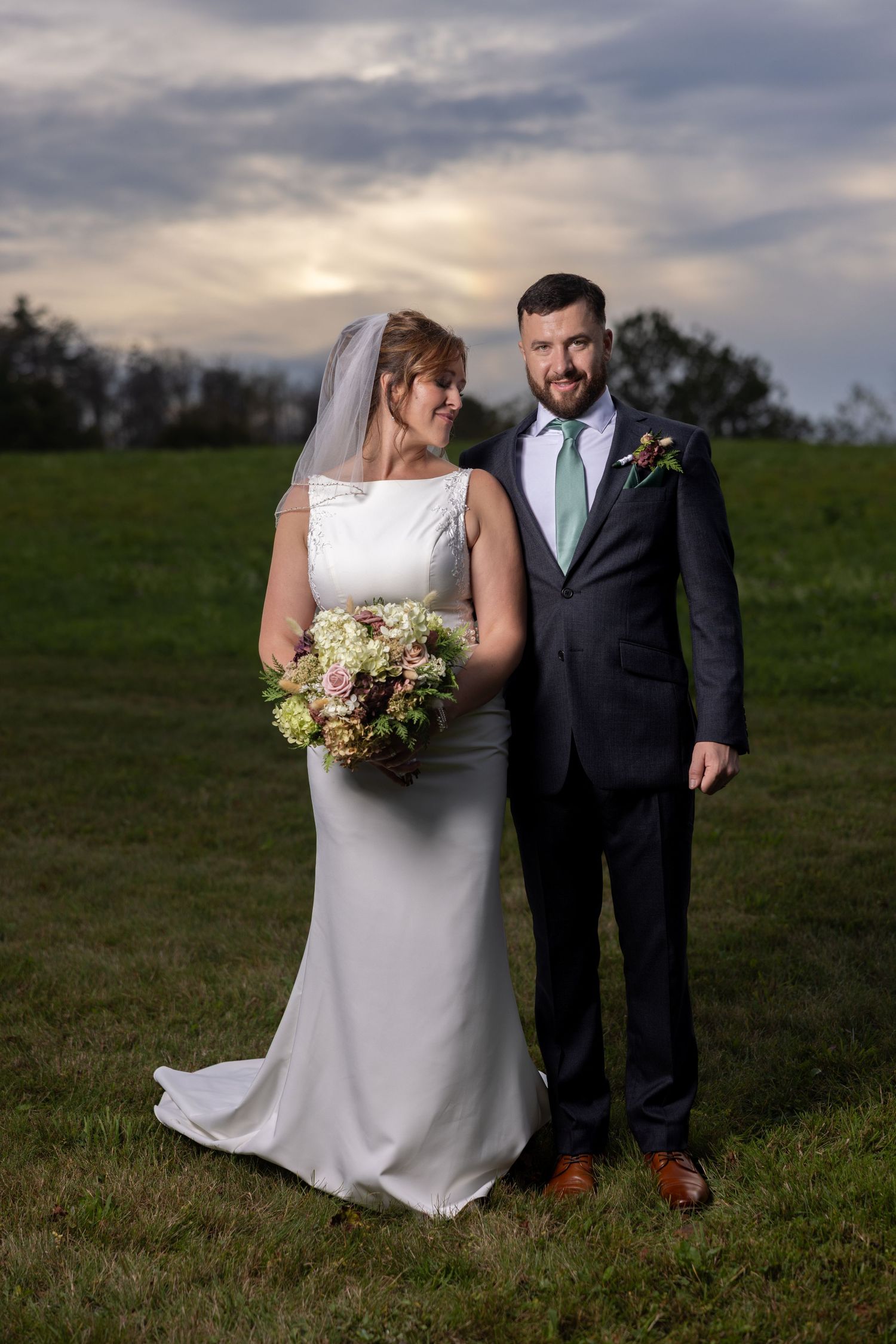 A bride and groom pose together on a grassy hillside at sunset, with the bride holding a pastel floral bouquet.