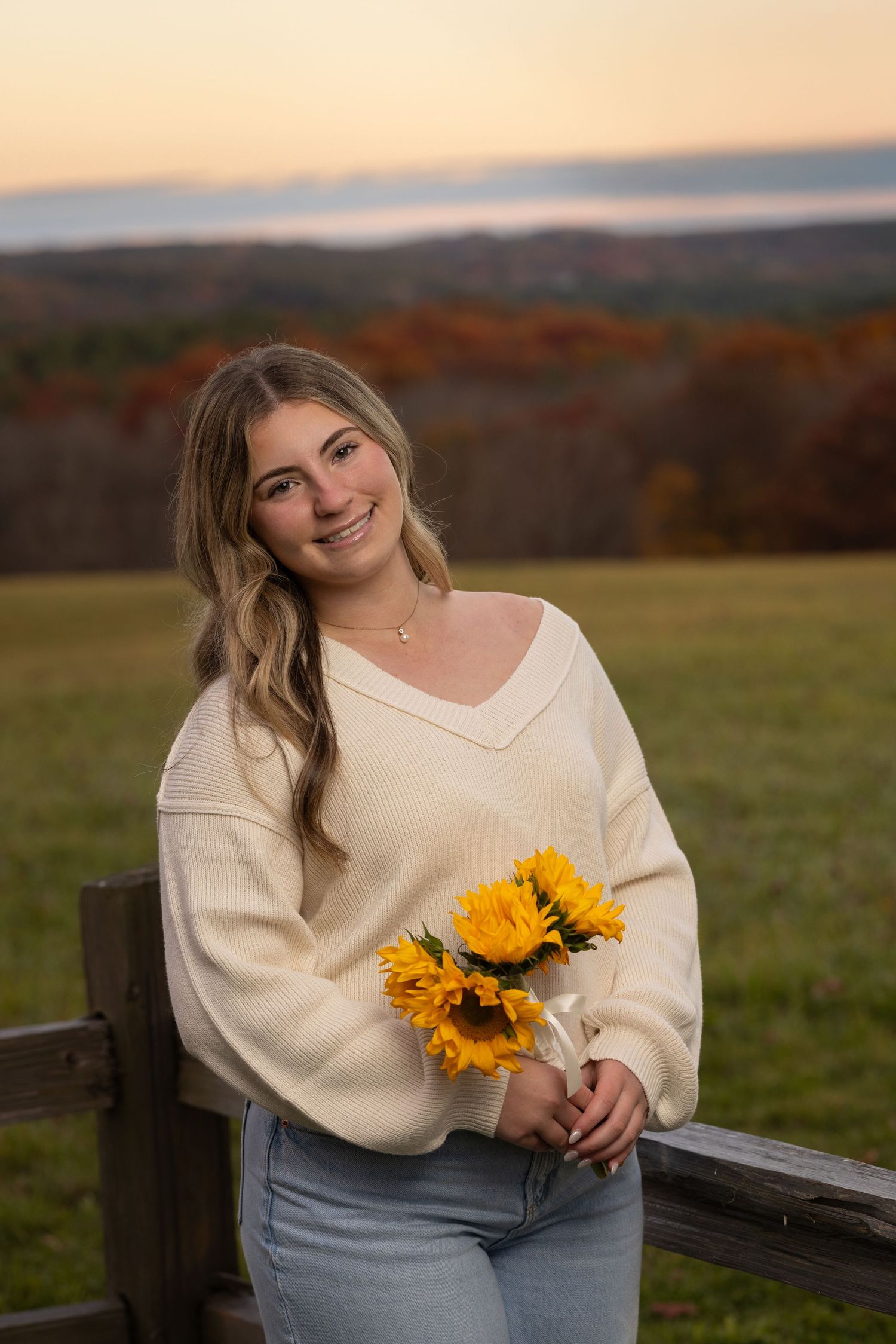Person in cream sweater holds sunflowers during autumn sunset photo session.