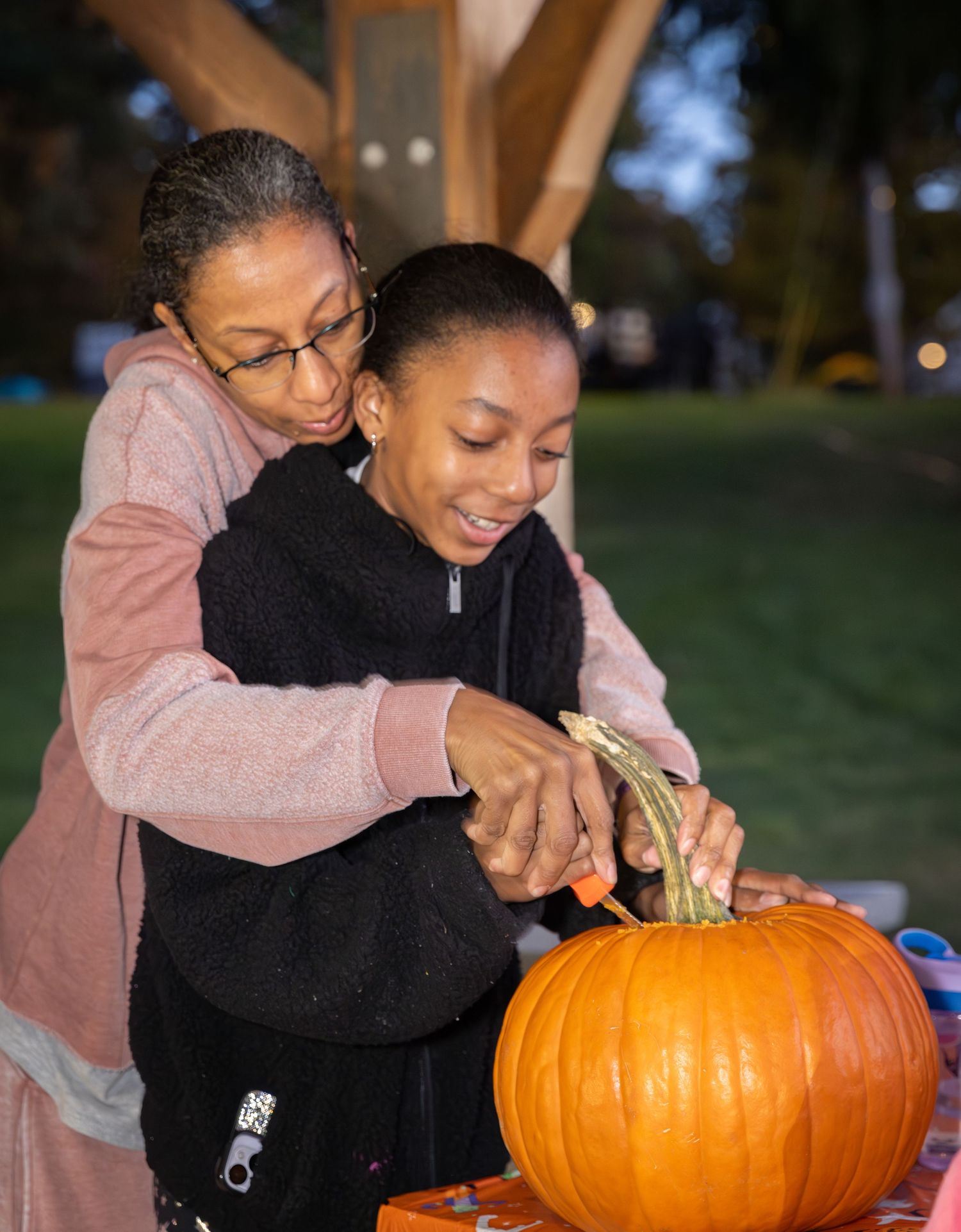 Two people carve a large orange pumpkin together outdoors on an autumn evening.