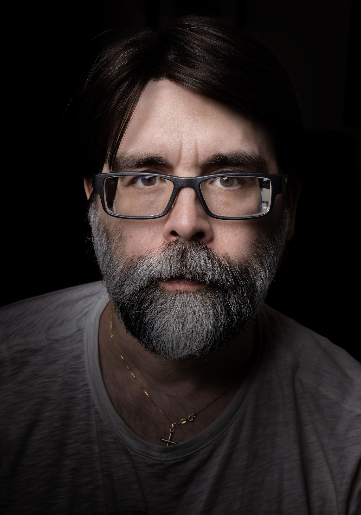 Low key portrait of a bearded person wearing glasses and a gray t-shirt against a dark background.