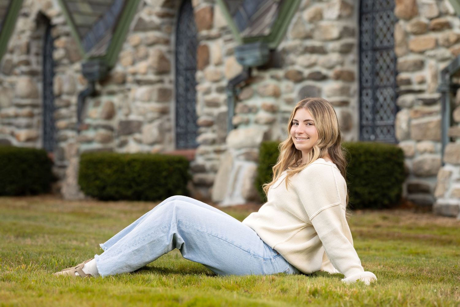 A person in a cream sweater and light jeans lies casually on grass near a stone church wall.