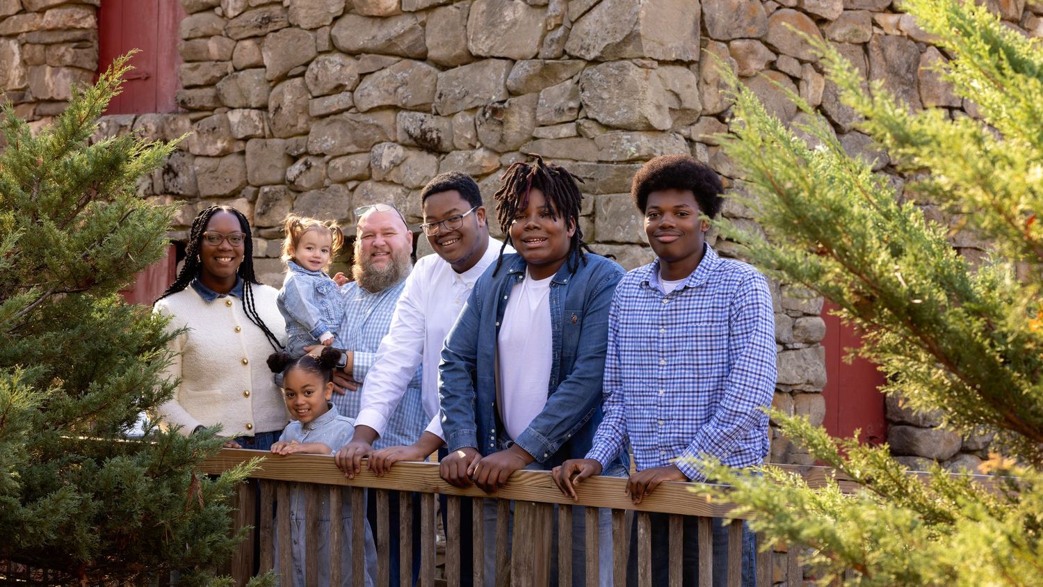 A group of people stand together smiling outdoors against a rustic stone wall backdrop in natural lighting.