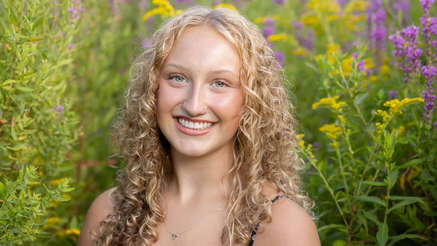 A person with curly blonde hair poses in a garden with vibrant purple and yellow wildflowers in the background.