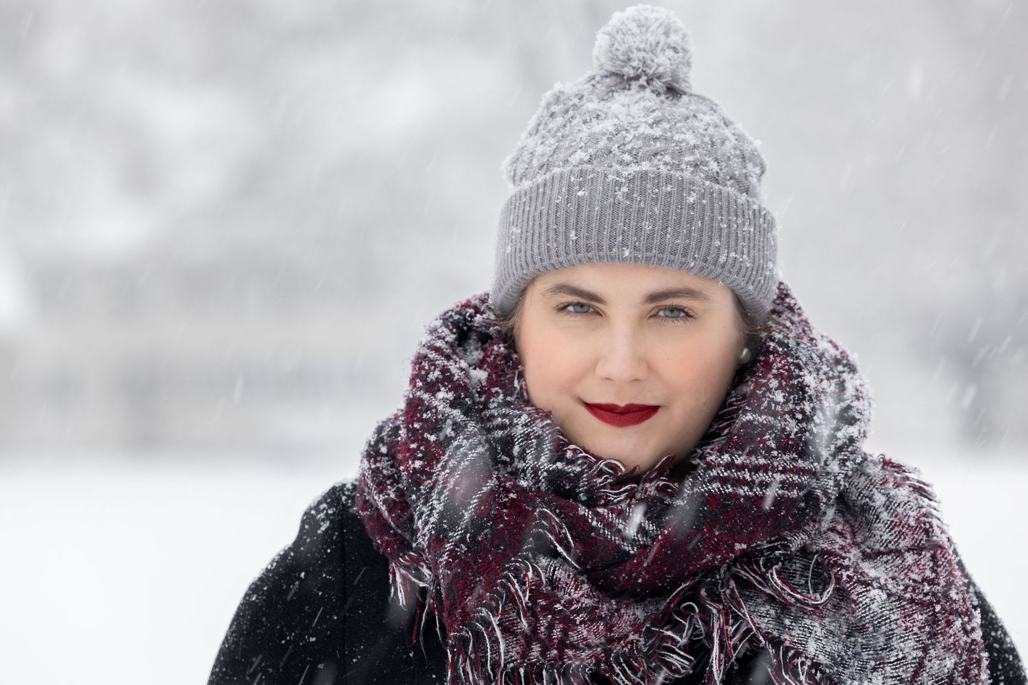 A person wearing a gray knit hat and scarf smiles during a snowy winter day with snowflakes falling around them.
