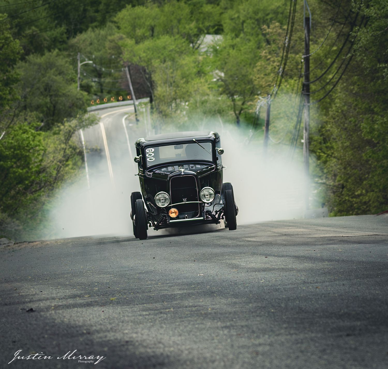 A black hot rod performs a burnout on a curved road, creating dramatic tire smoke against a green forest backdrop.