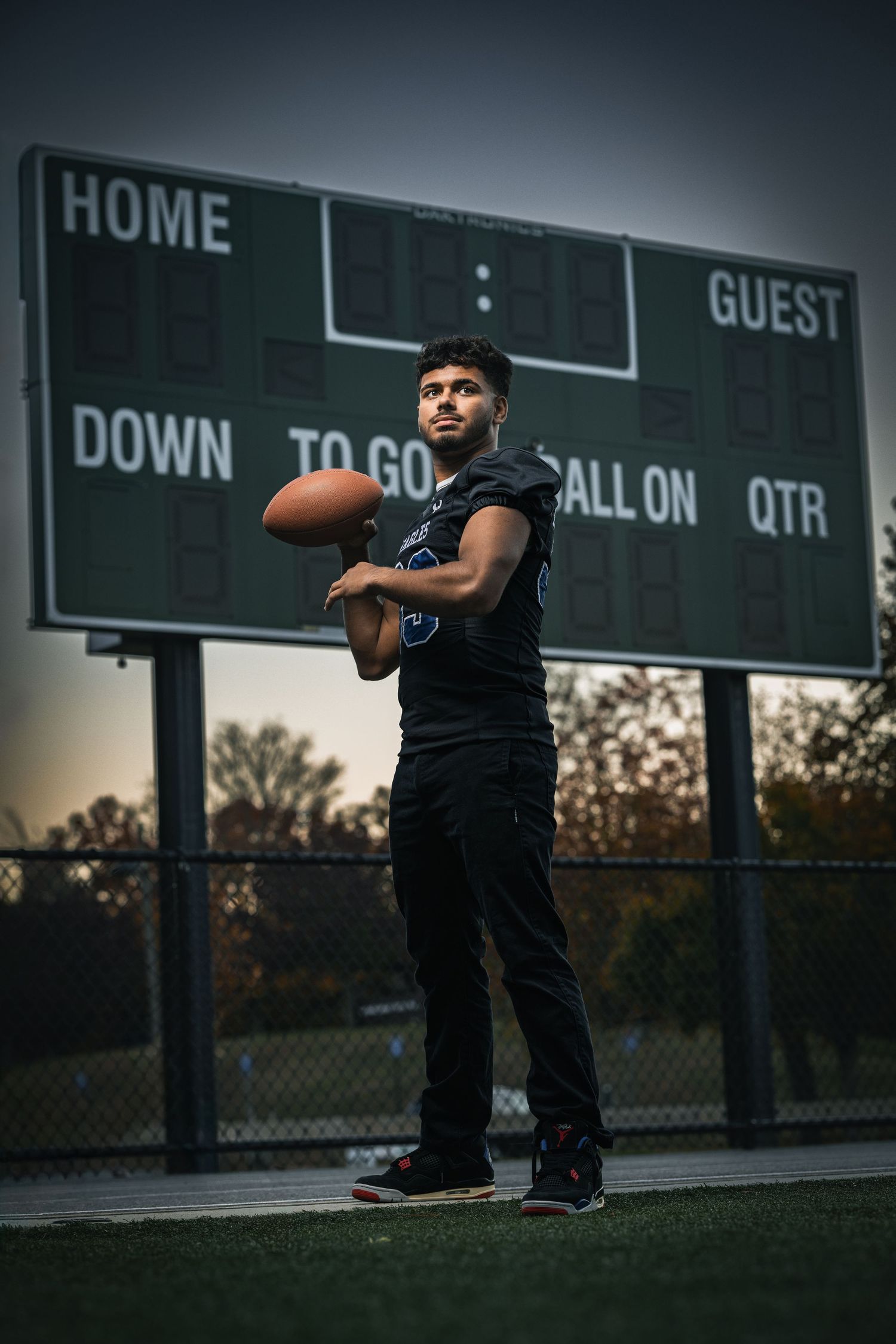 A football player poses with a ball on a field near a scoreboard at dusk.