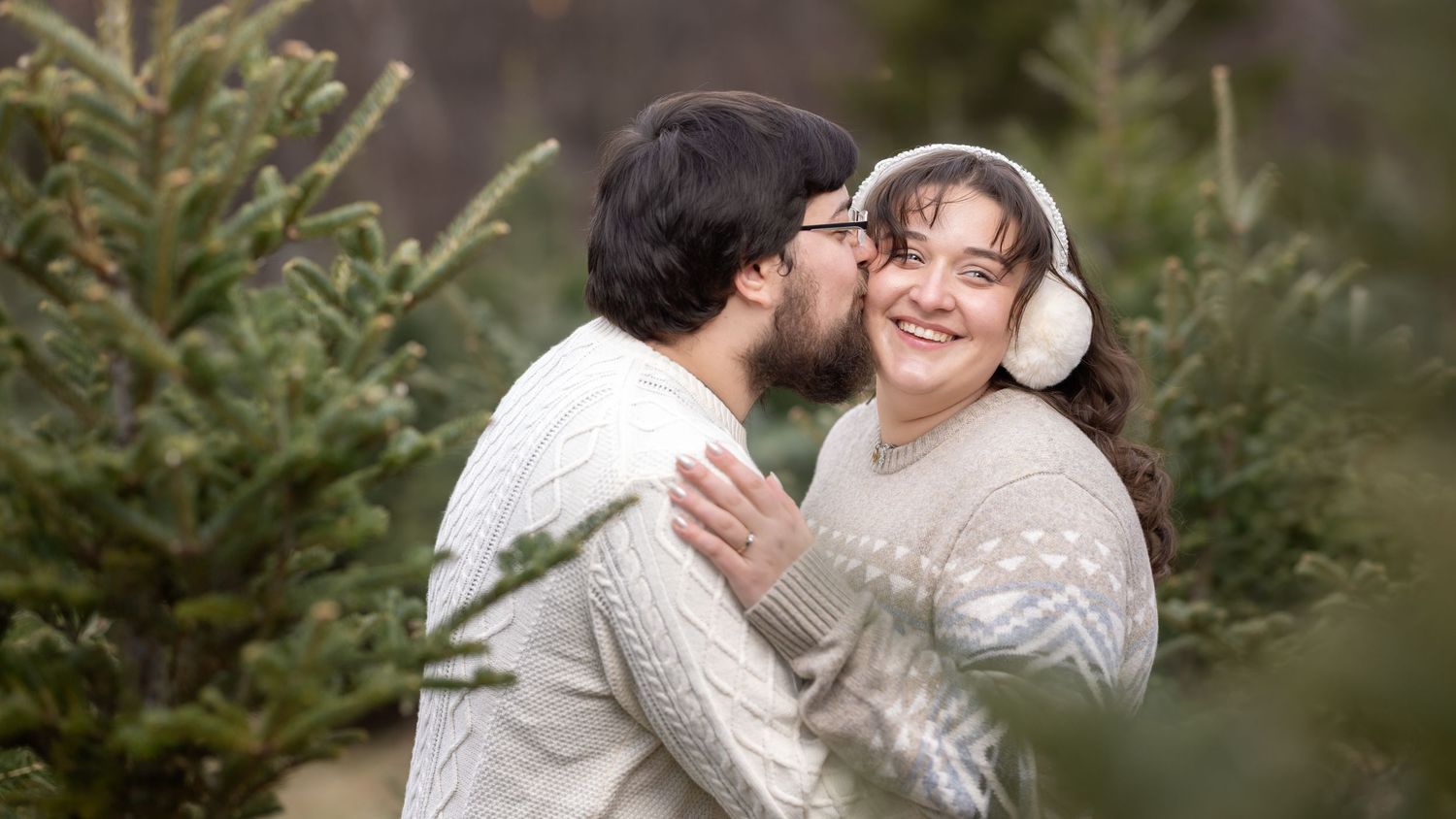 A couple in cozy white sweaters share a tender moment among evergreen trees in a winter setting.