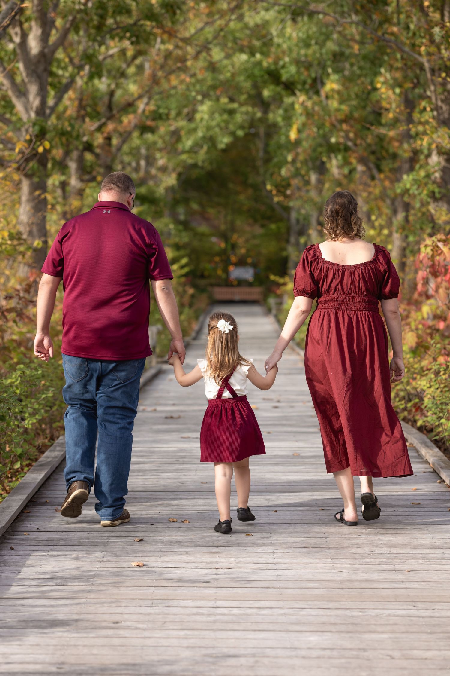 Family wearing matching burgundy outfits walks hand in hand down a wooden boardwalk path surrounded by autumn trees.