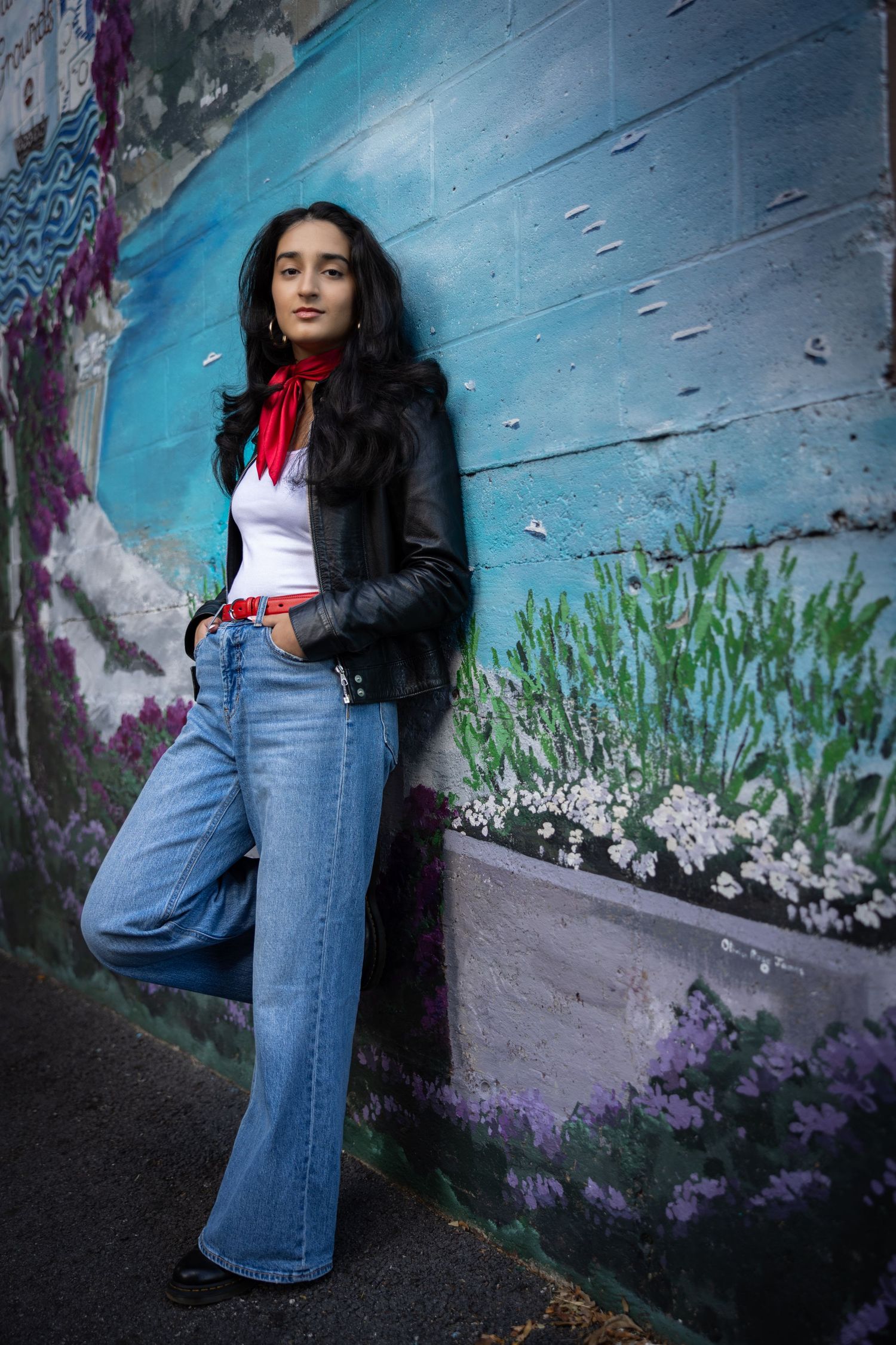 A person in flared jeans and leather jacket poses against a blue wall with street art and flowers.