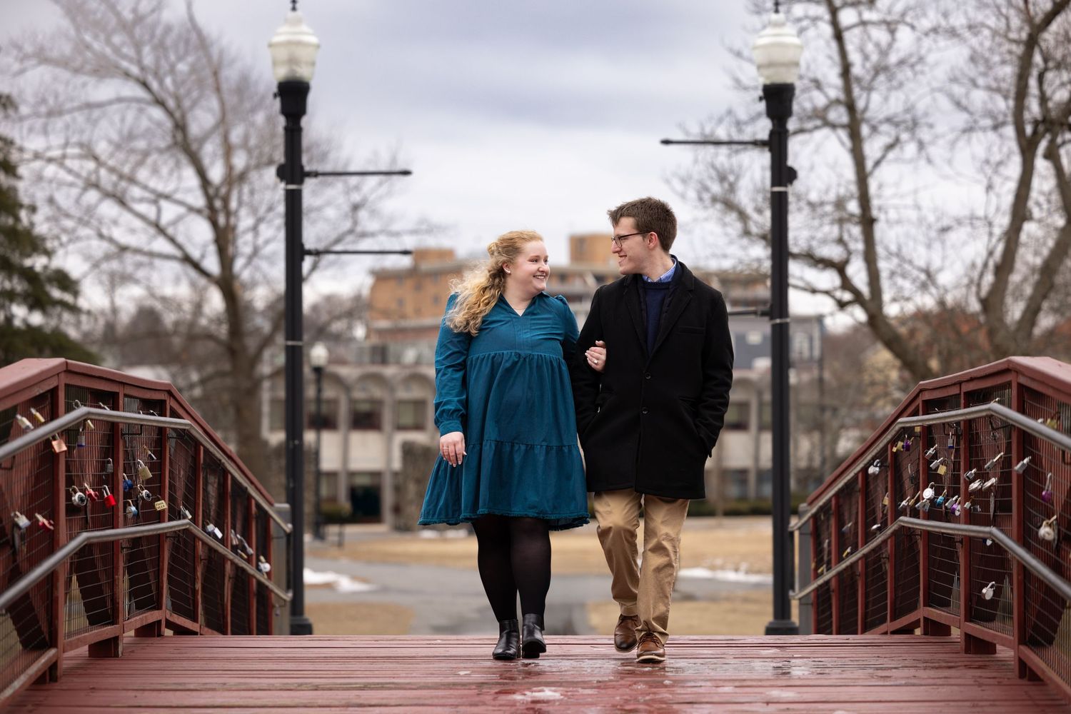 A couple in blue and black winter coats share a romantic moment while walking across a rustic wooden bridge.