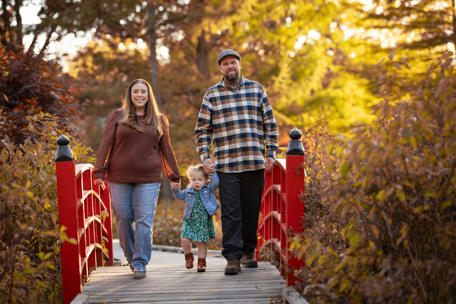 A family walks together on a red wooden bridge surrounded by golden autumn foliage during sunset.
