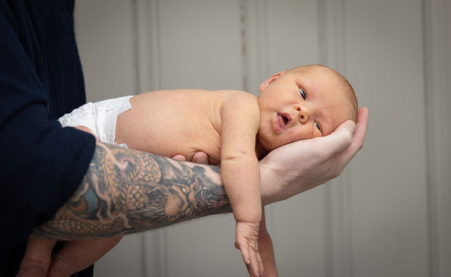 A tiny newborn baby rests peacefully on a tattooed arm against a light background.