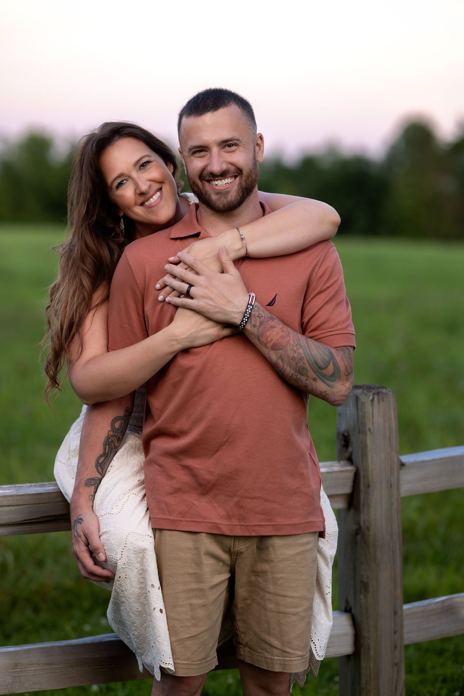 A couple shares joyful laughter while sitting together on a wooden fence in a green field at sunset.