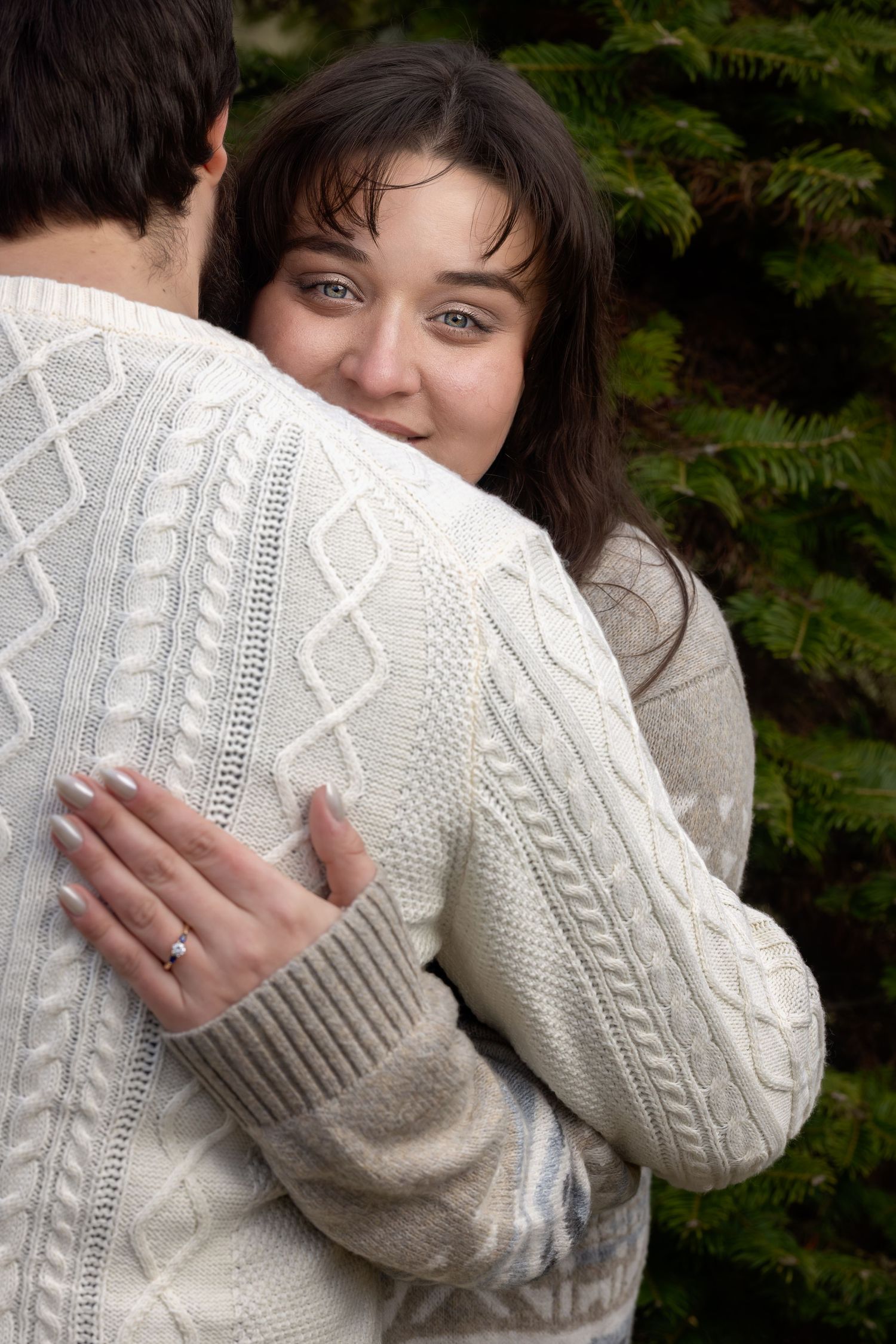 A couple embraces while wearing a cozy white cable knit sweater against an evergreen background.