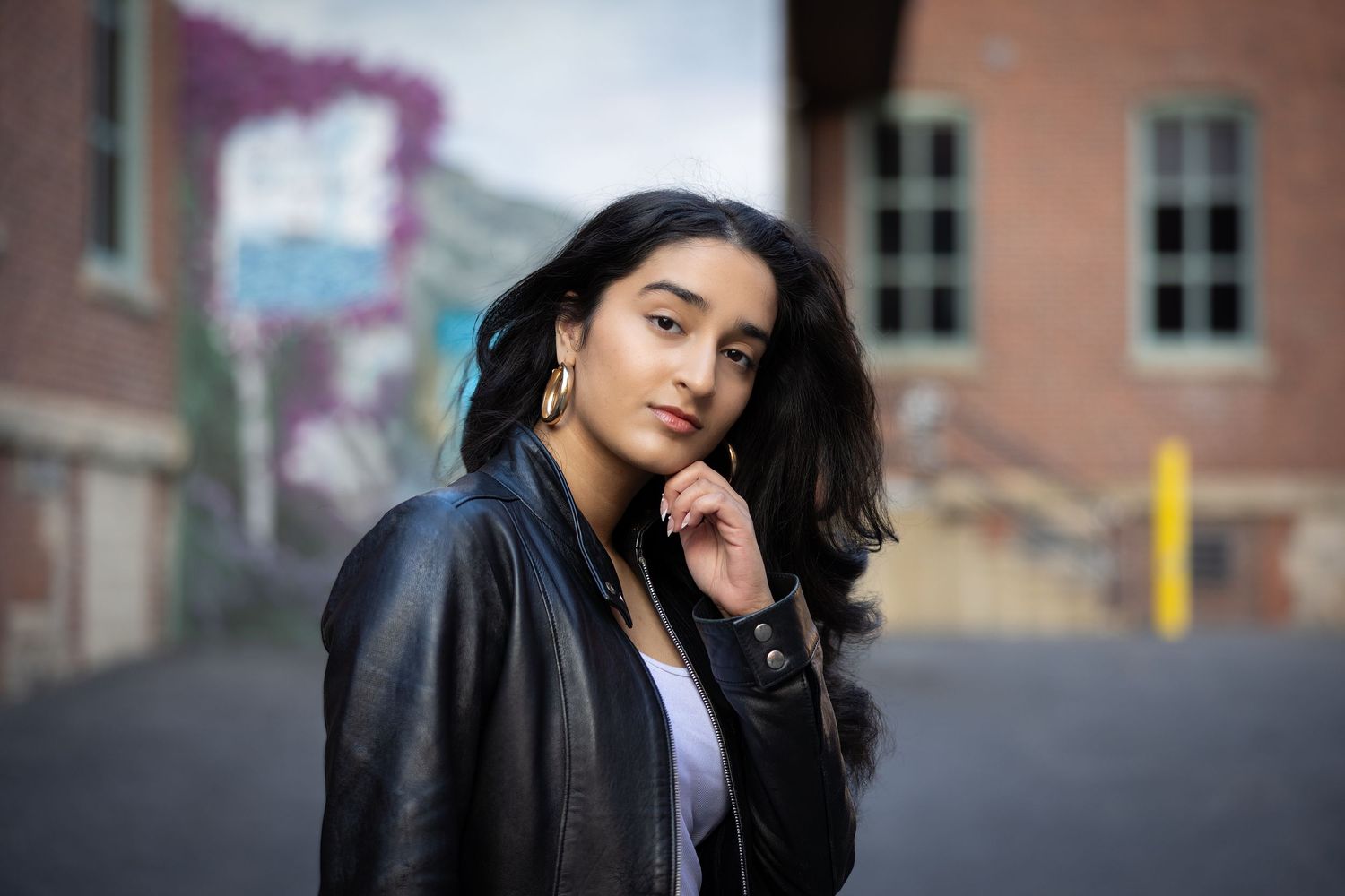 A person with long dark hair wearing a black leather jacket poses against a blurred brick building background.