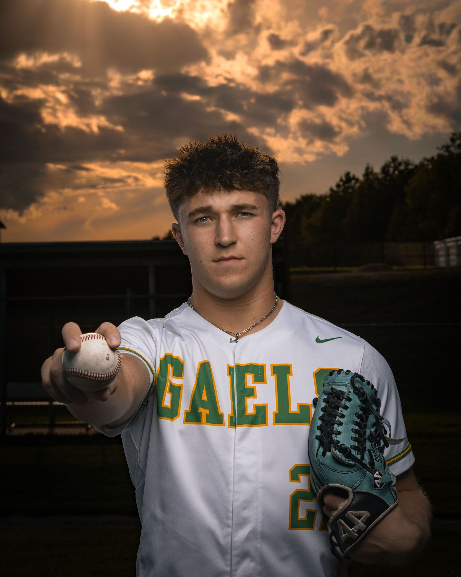 Baseball player in white Valley uniform poses with glove and ball against dramatic sunset sky.