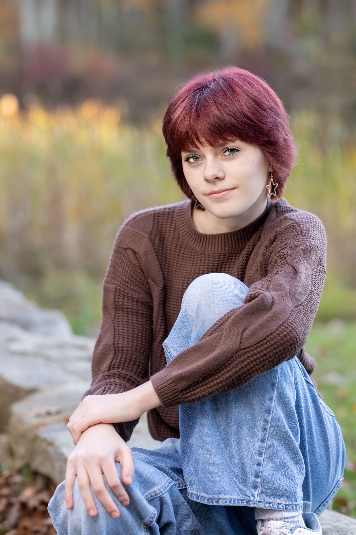 A person in a brown knit sweater and blue jeans sits thoughtfully on the ground in an autumn outdoor setting.