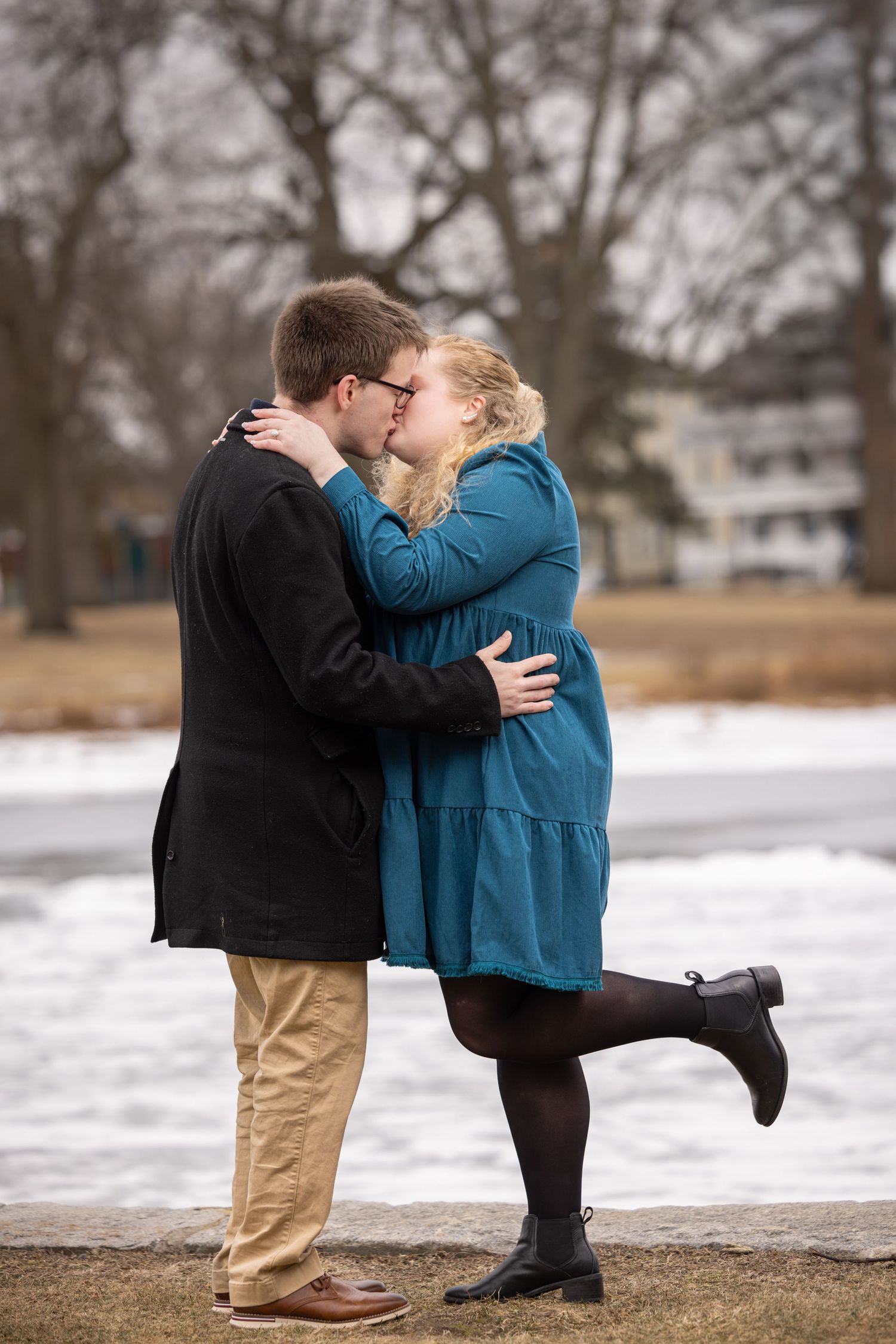 A couple embraces and kisses in a winter landscape with snow on the ground and trees in the background.