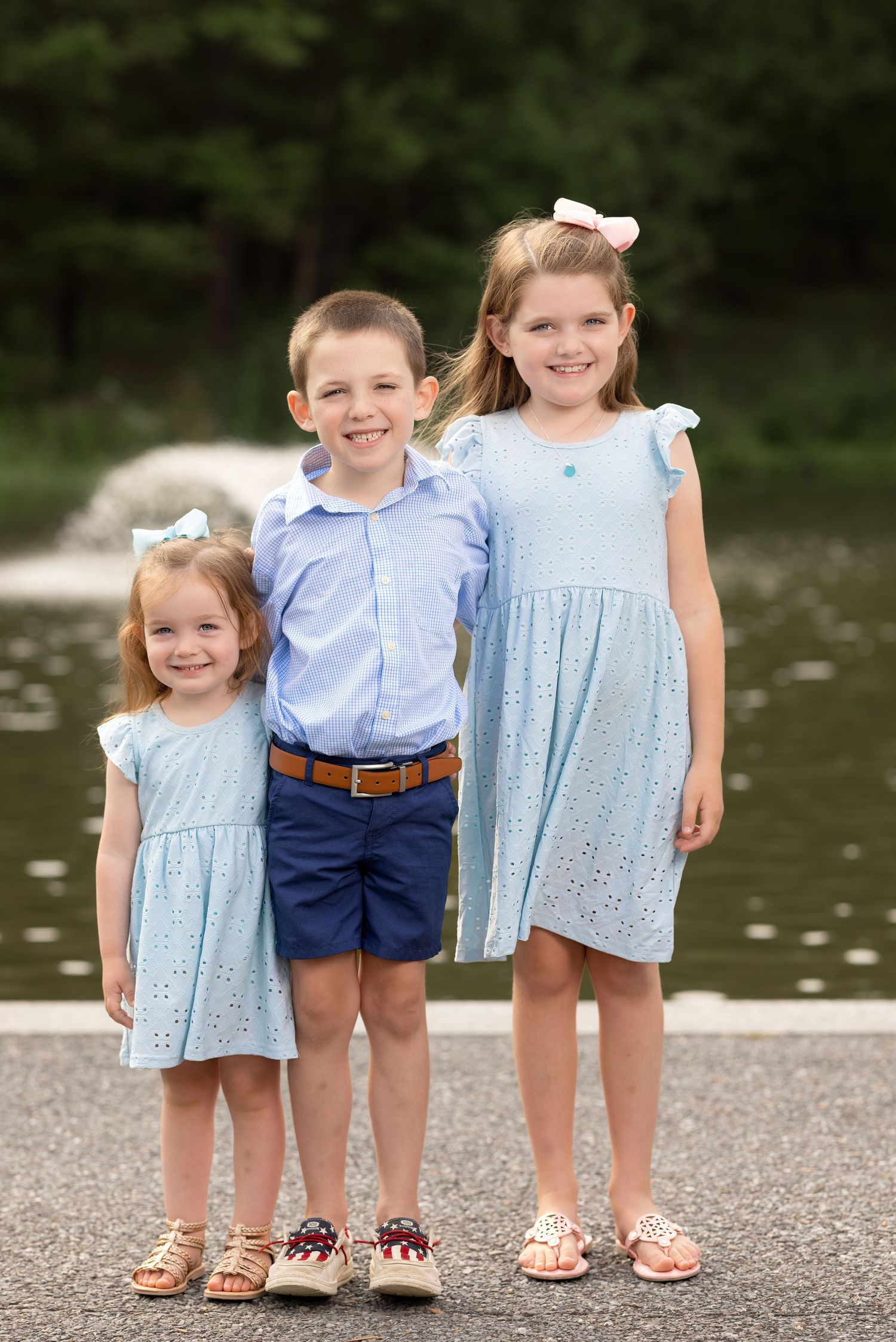 Three children wearing coordinated blue and white outfits stand together on a gravel path near water.