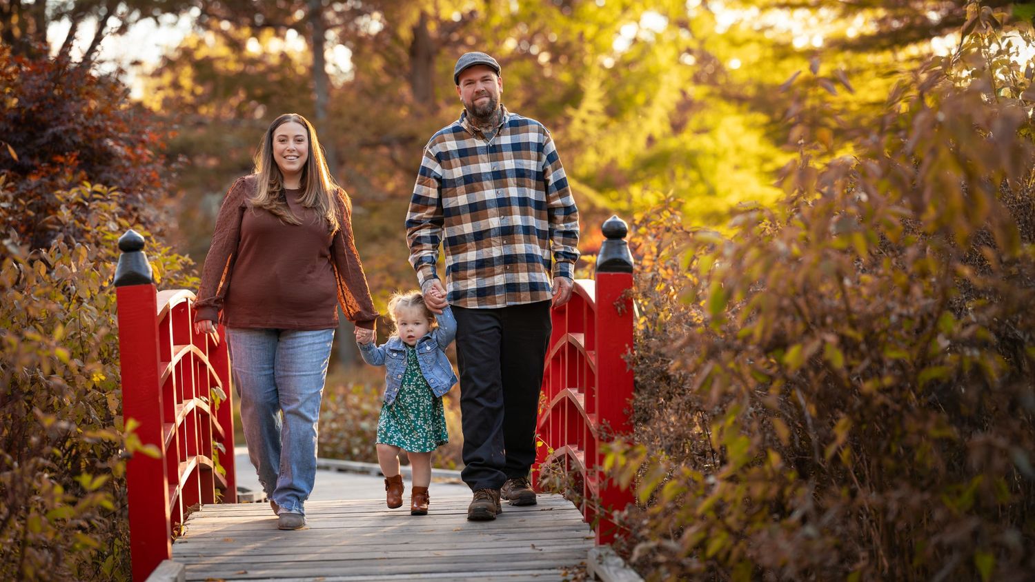 Family walking together on a red wooden bridge surrounded by golden autumn foliage at sunset.