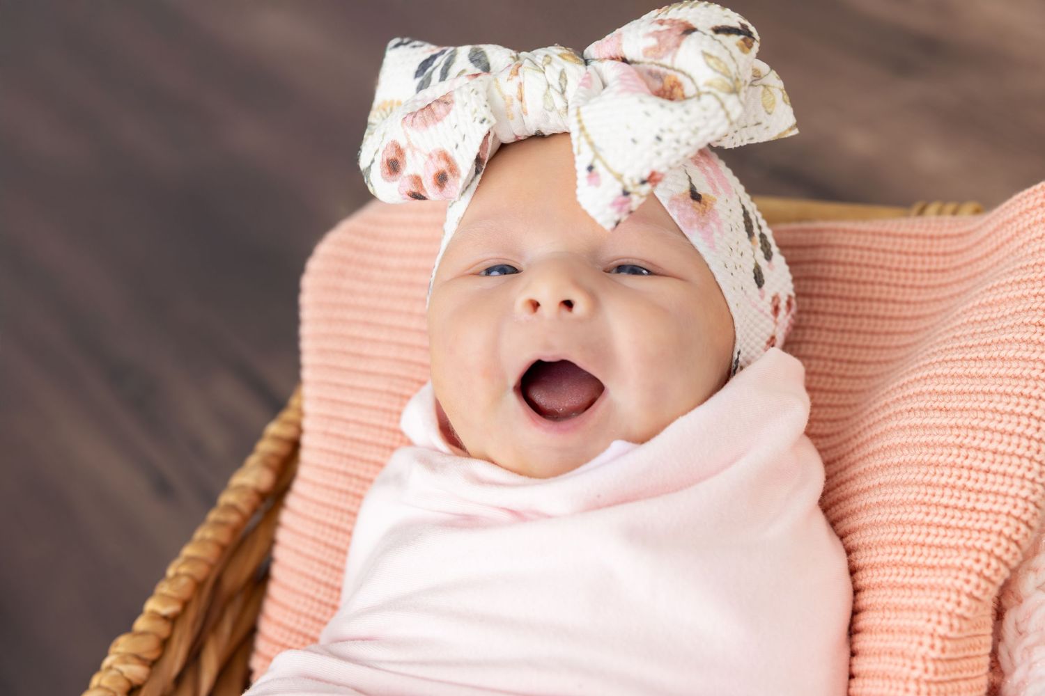 Baby wearing pink clothing and floral headband smiles while lying on a coral-colored blanket.