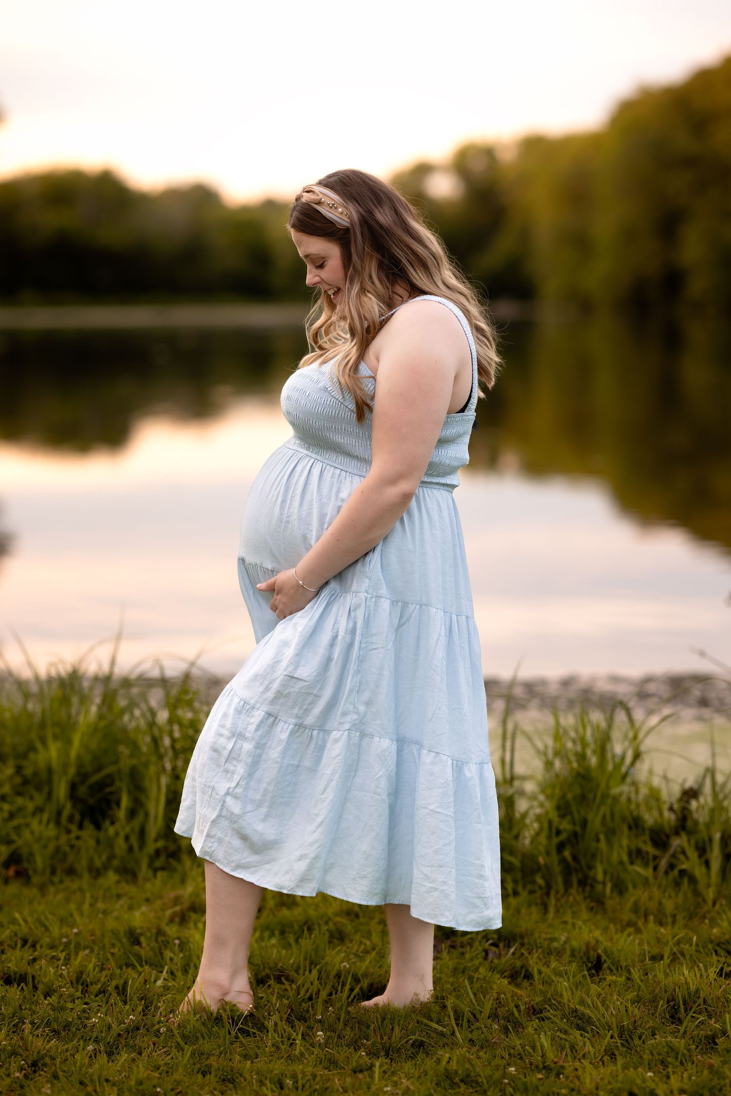 A person in a light blue dress bends down to pick wildflowers by a serene lake at sunset.
