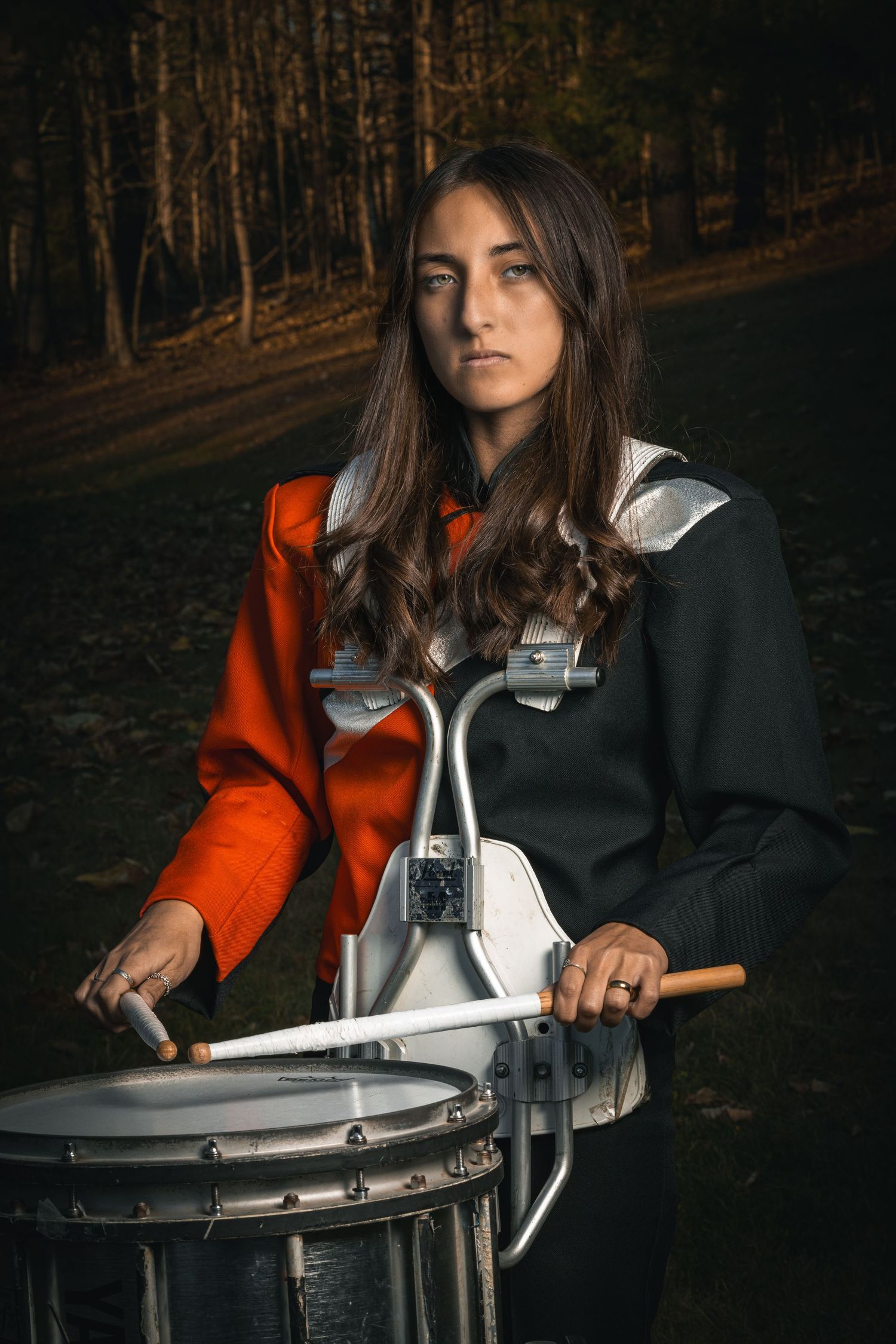A drummer in an orange jacket plays a marching band snare drum in a dark forest setting.