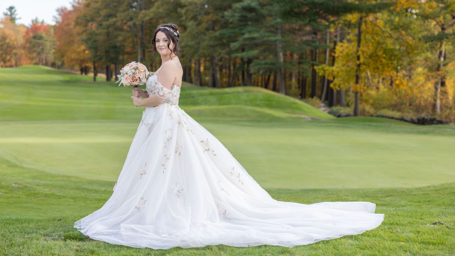 A person in a flowing white wedding dress poses gracefully on a lush green golf course surrounded by autumn trees.