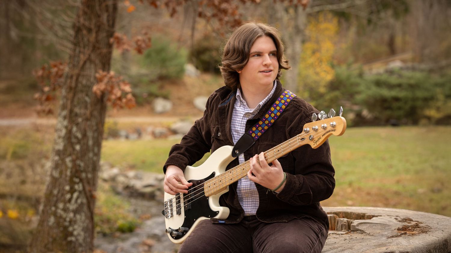Musician in dark attire plays guitar outdoors in autumn setting with fallen leaves and natural scenery.