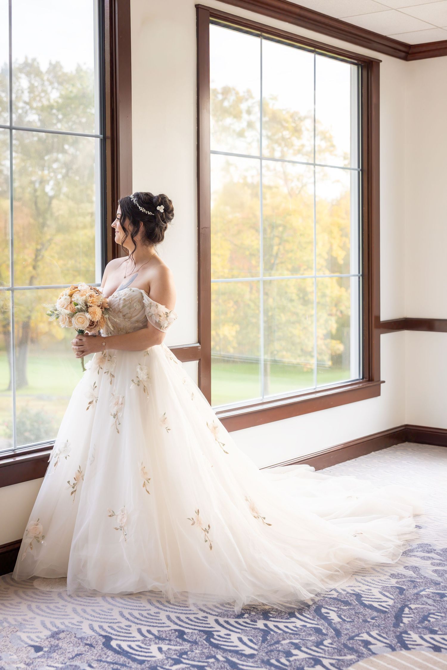 A bride in an elegant white ball gown with gold embellishments stands by a large window overlooking autumn trees.