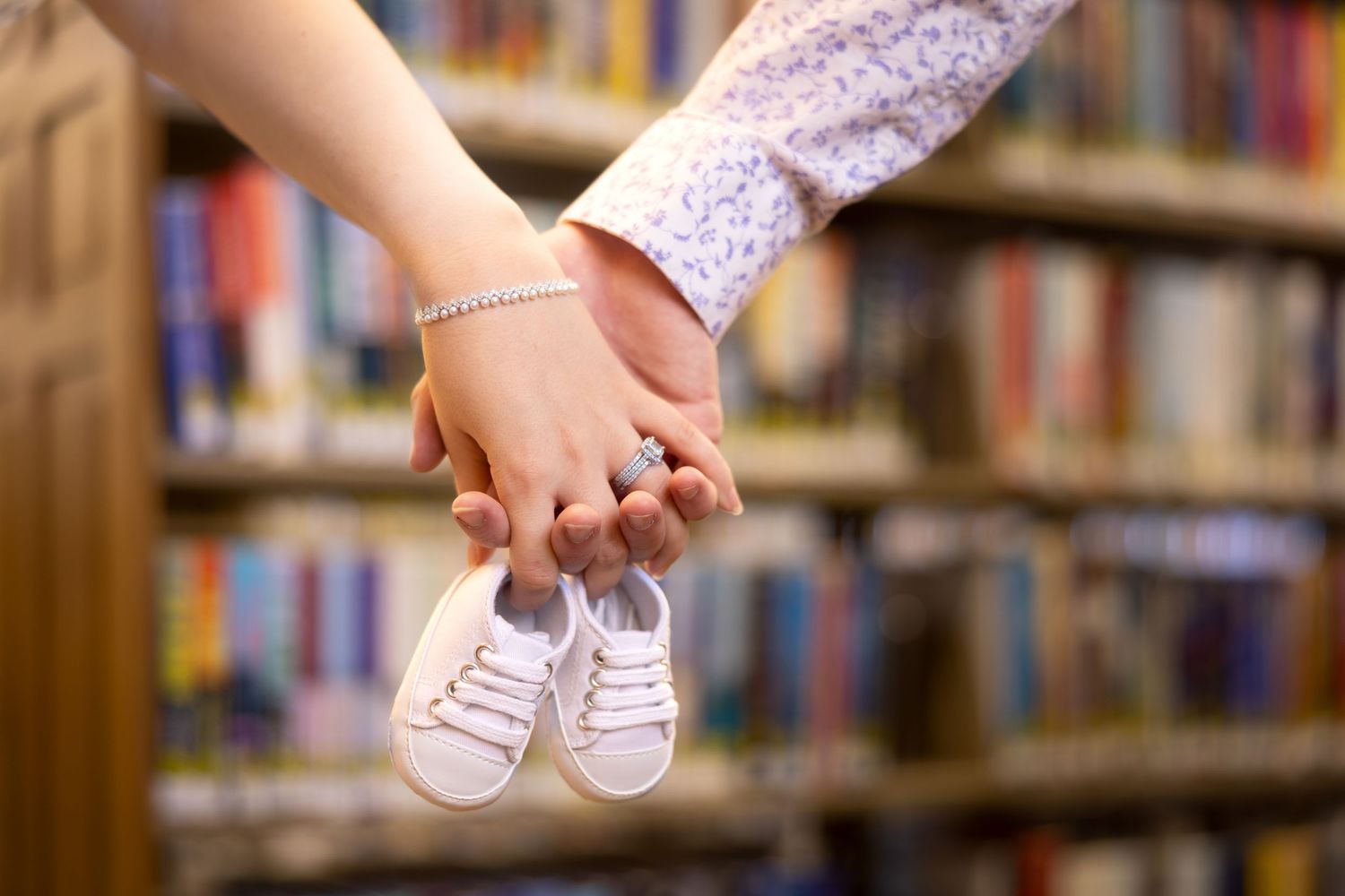 Small pink baby shoes being held up in a library with bookshelves visible in the background.