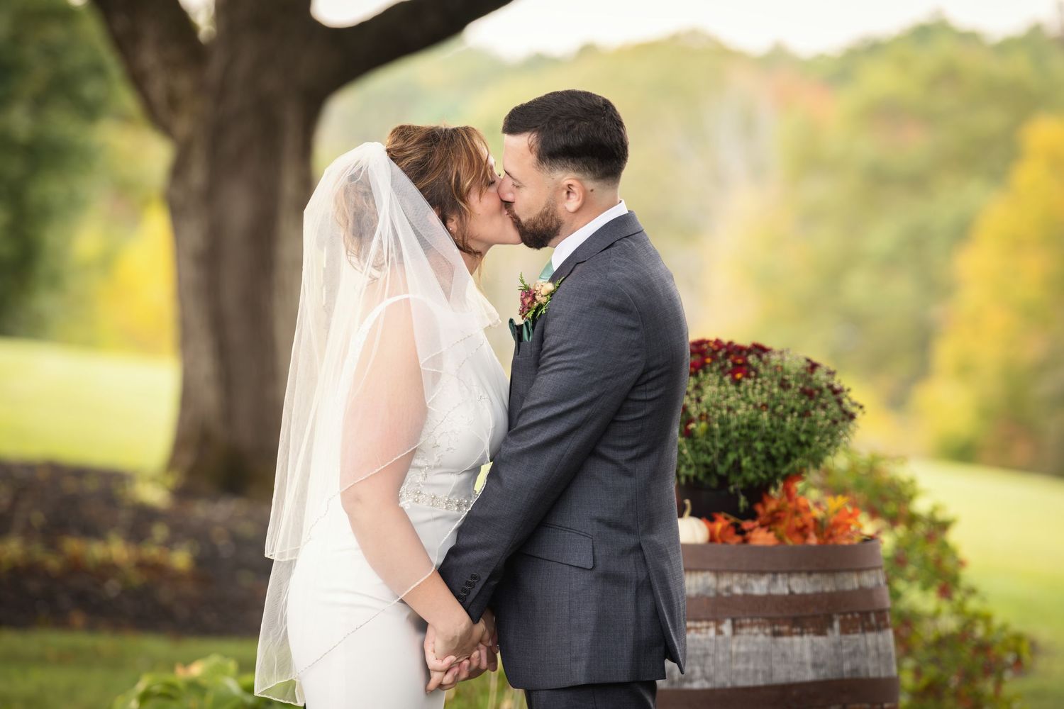 Newlywed couple embracing outdoors with autumn decorations and fall foliage in background.