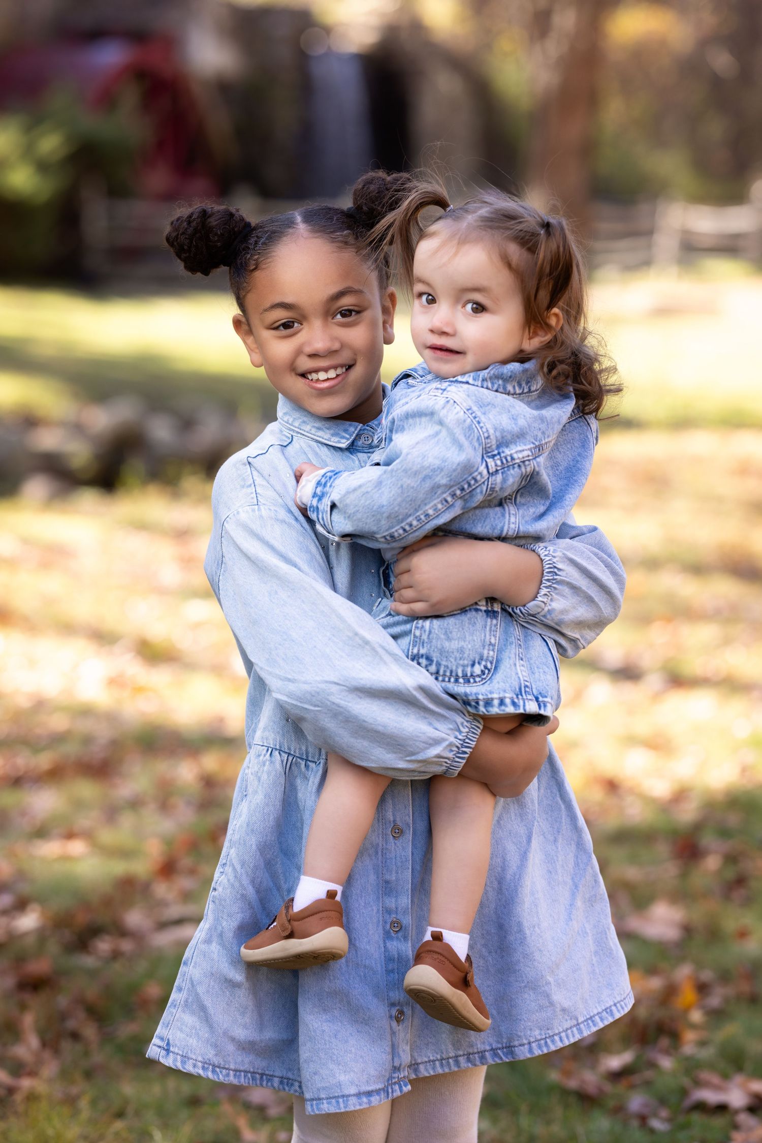 Two children in matching denim outfits hug and play together on fallen autumn leaves in a park.