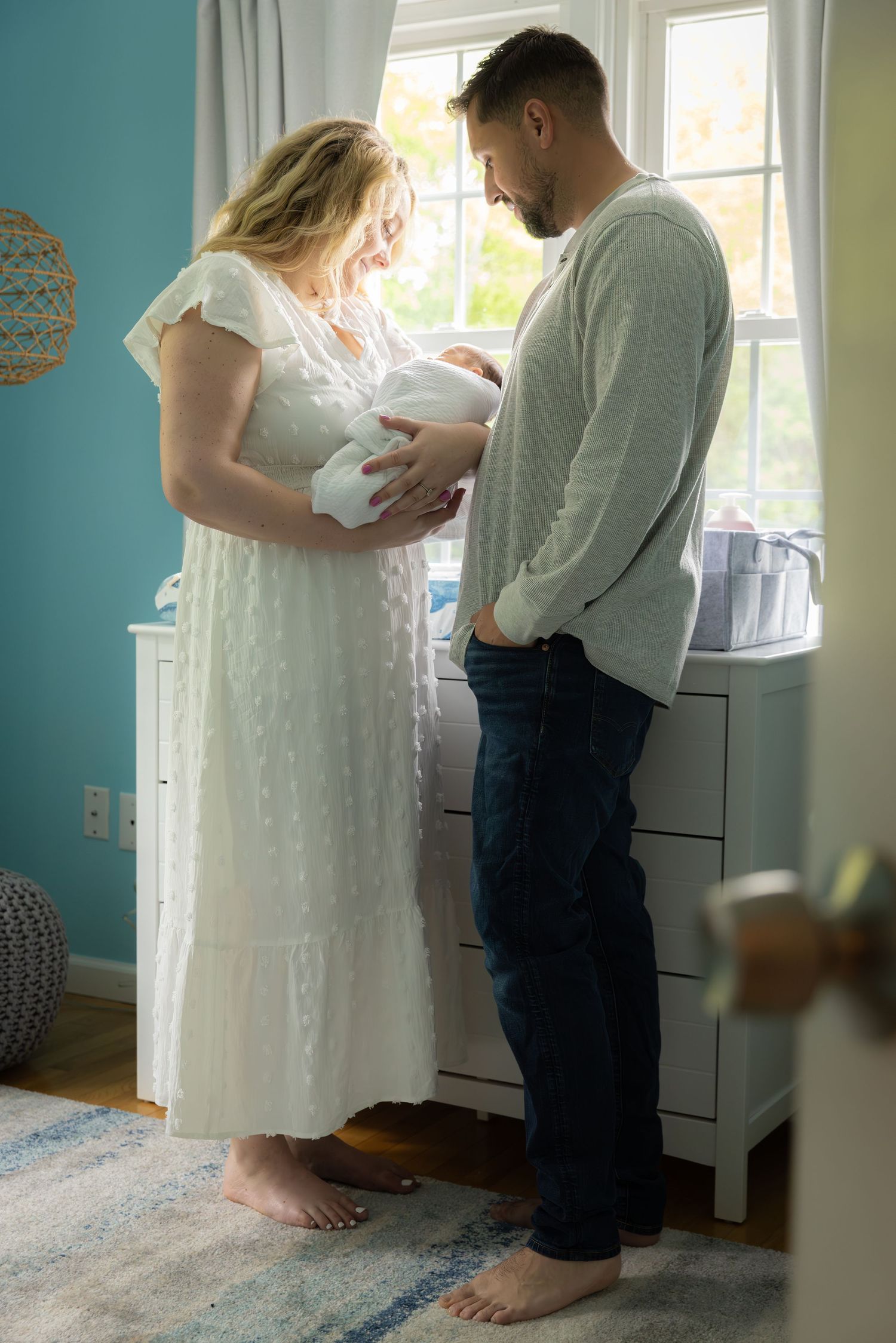 Parent and child stand together in a bright nursery room with turquoise walls and white curtains.