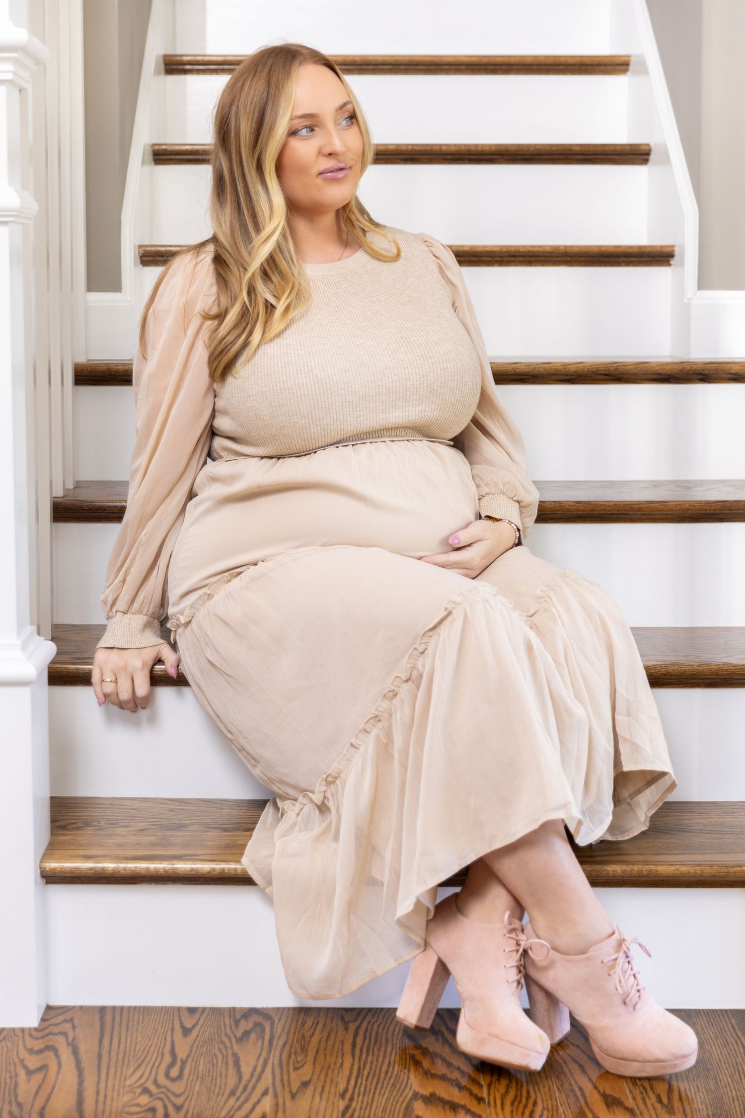 Someone in a flowing beige dress sits on wooden stairs while wearing cozy pink slippers.