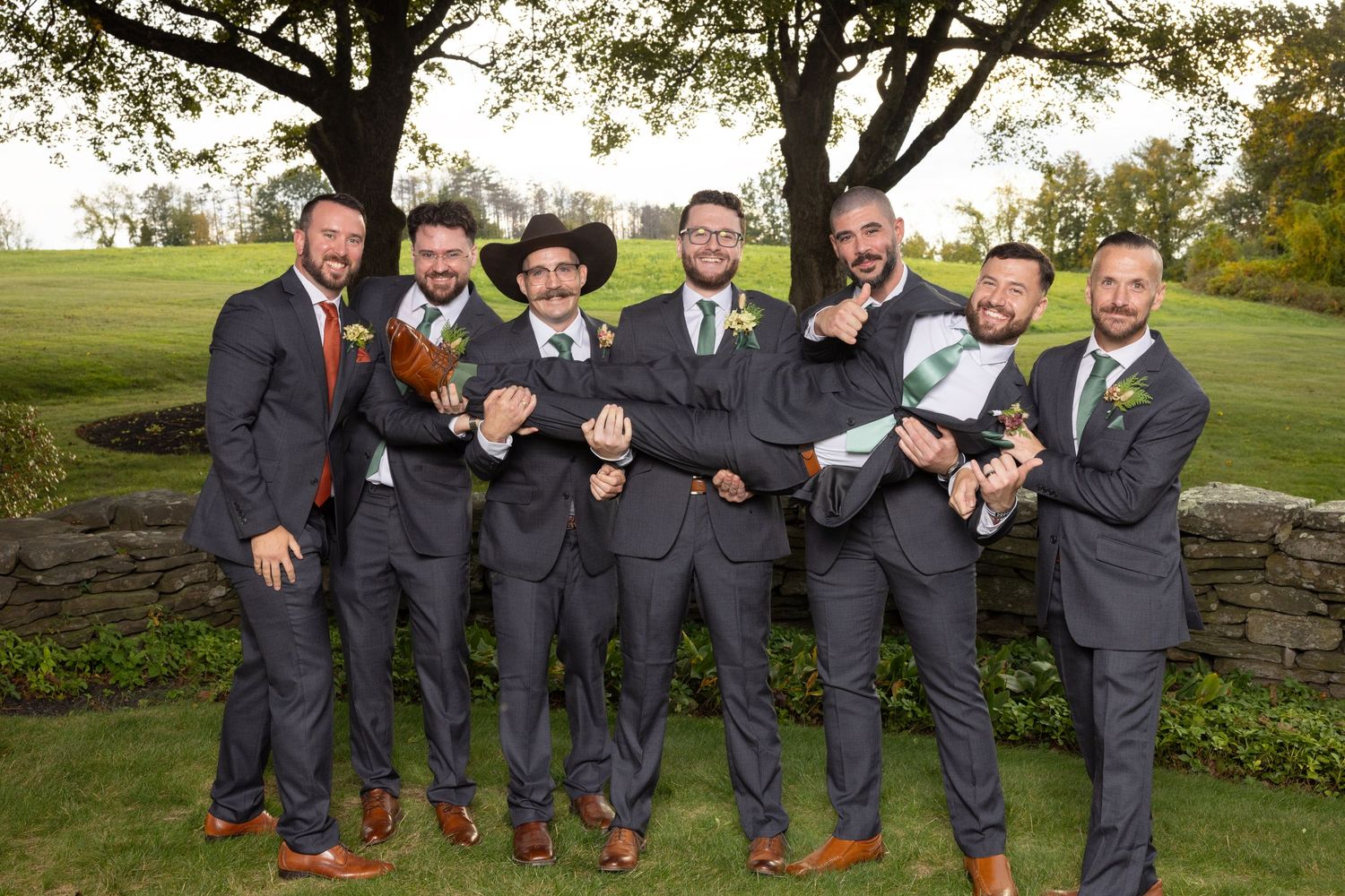 Groomsmen in charcoal suits sit casually on a stone wall outdoors during a wedding photoshoot.