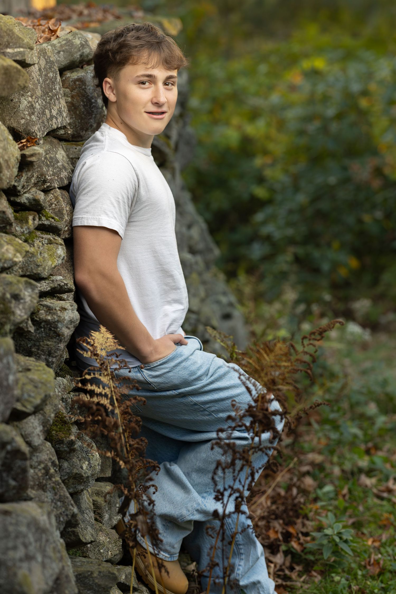 A person in a white t-shirt and jeans sits casually on a stone wall surrounded by autumn foliage.